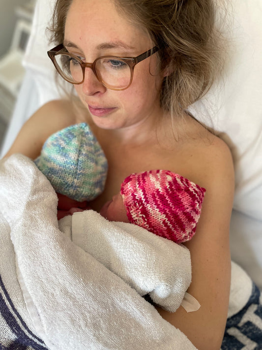 Mother staring forlorn into the distance, holding newborn twins on her bare chest, on a hospital bed.