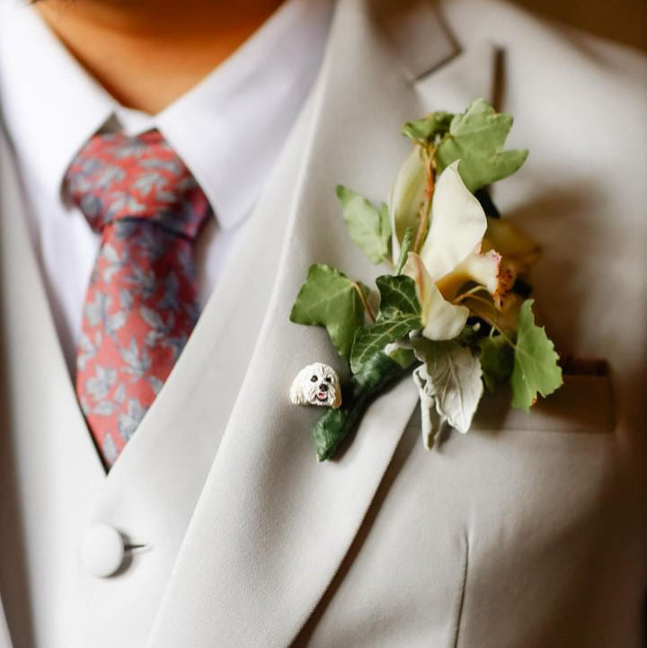 A custom pet lapel pin in the shape of a dog, attached to the lapel of a suit jacket, next to a floral boutonnière.
