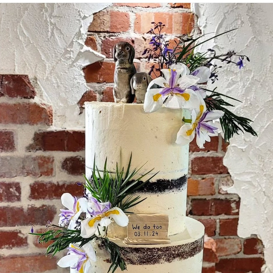 Decorative wedding cake with flowers and text, against a brick wall background, featuring custom pet rabbit cake toppers.