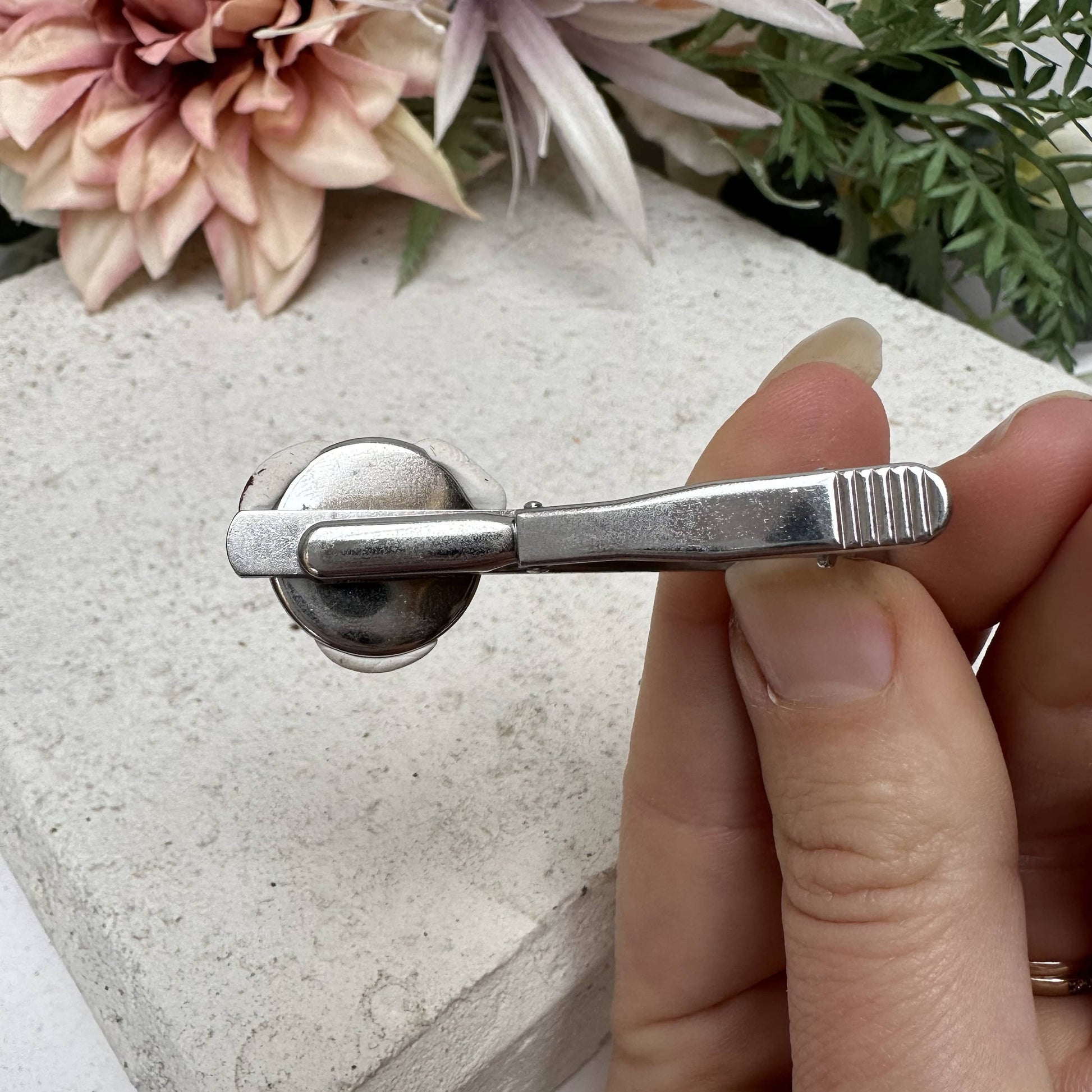 Silver tie clip held by a hand with flowers and greenery in the background