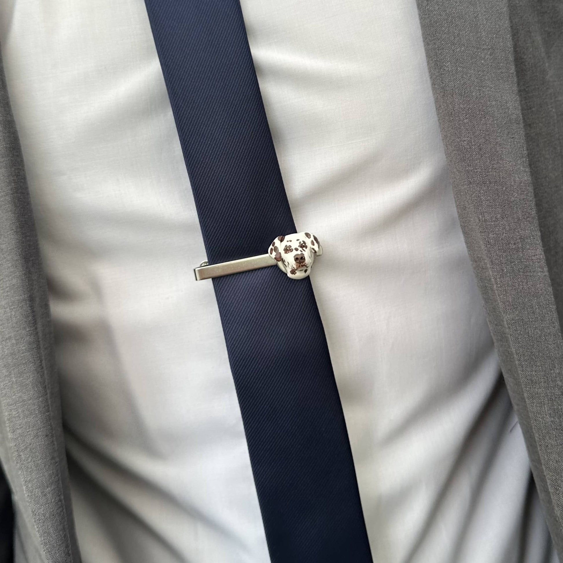 Close-up of a navy tie with a silver tie clip worn by a person in a gray suit. Tie clip has a custom pet face feature.