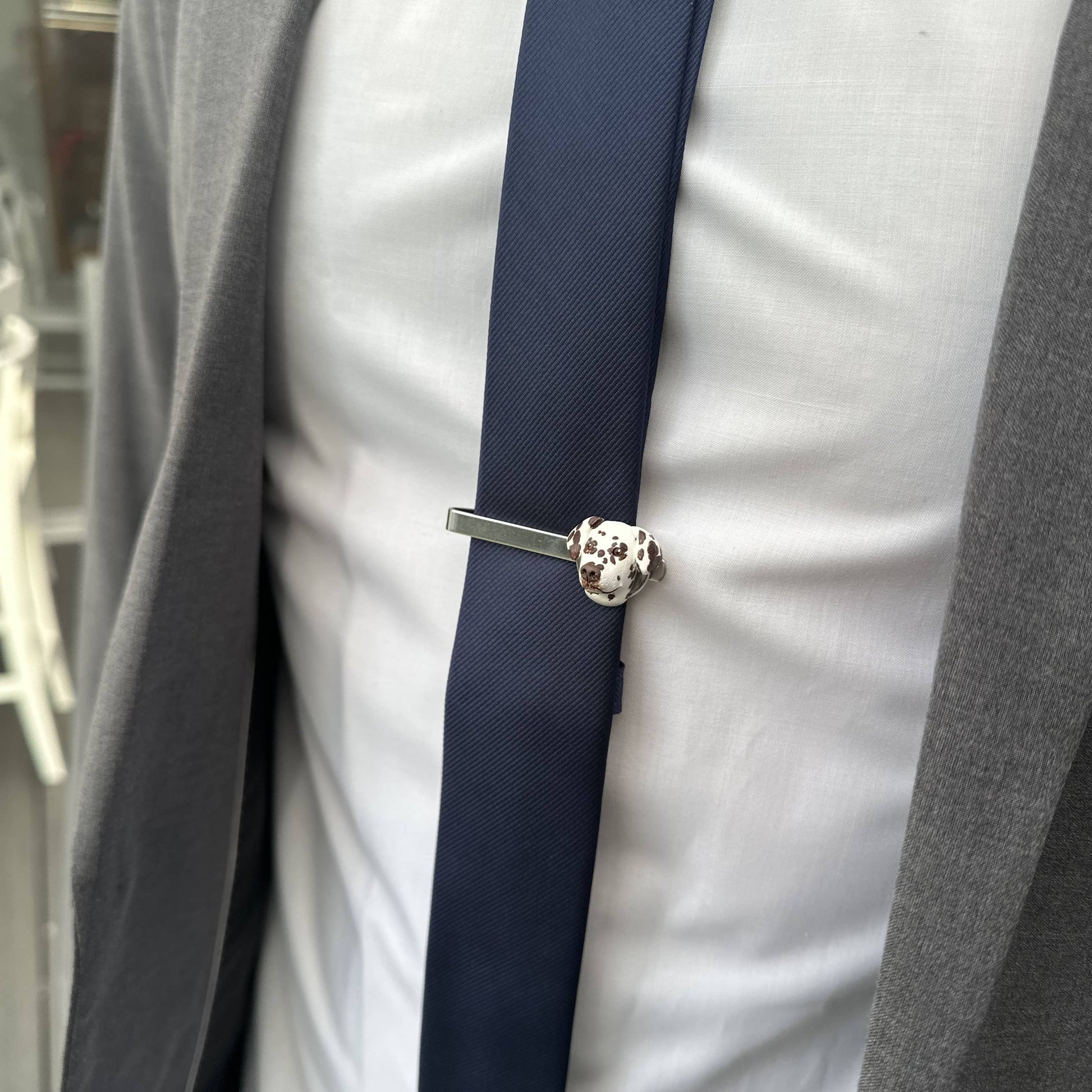 Close-up of a silver tie clip on a navy blue tie against a gray suit background. Tie clip has a custom pet face feature.
