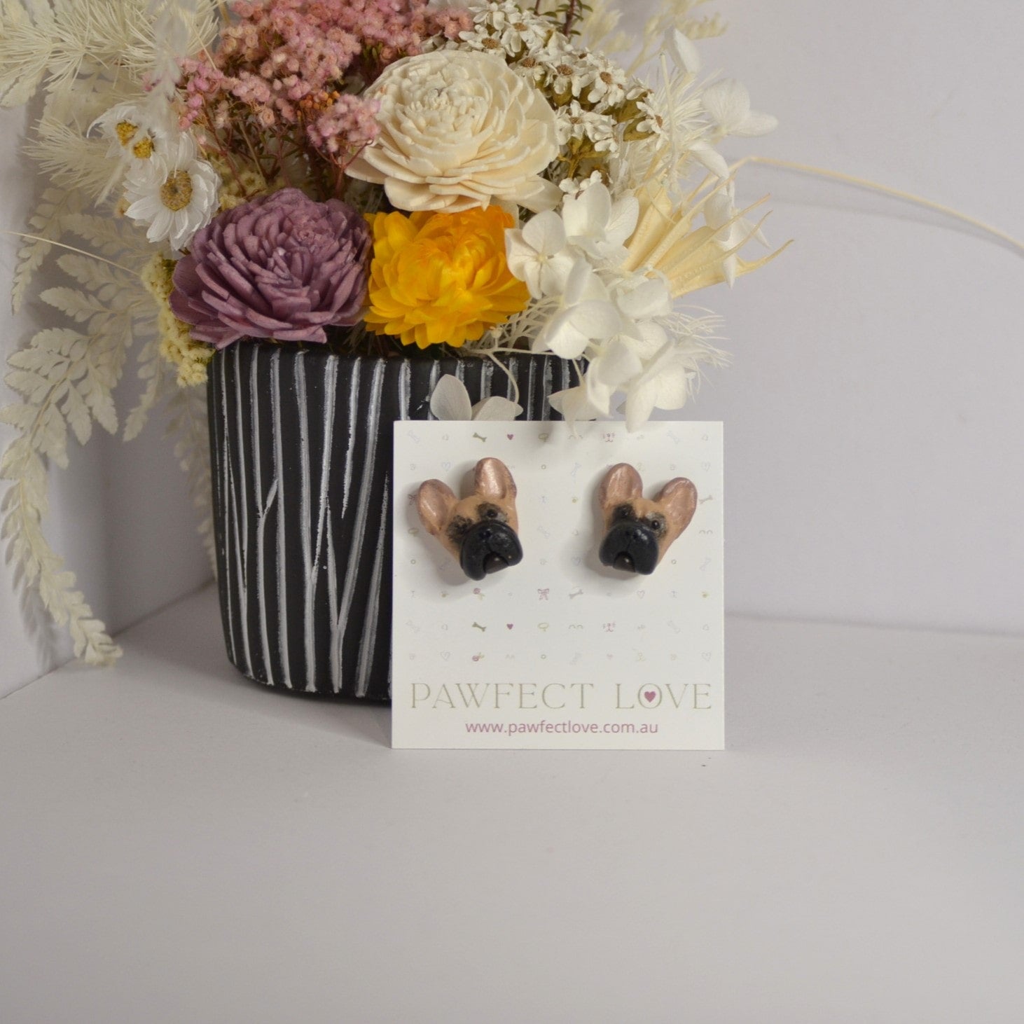 Handmade French bulldog stud earrings by Pawfect Love, positioned in front of green flower arrangement