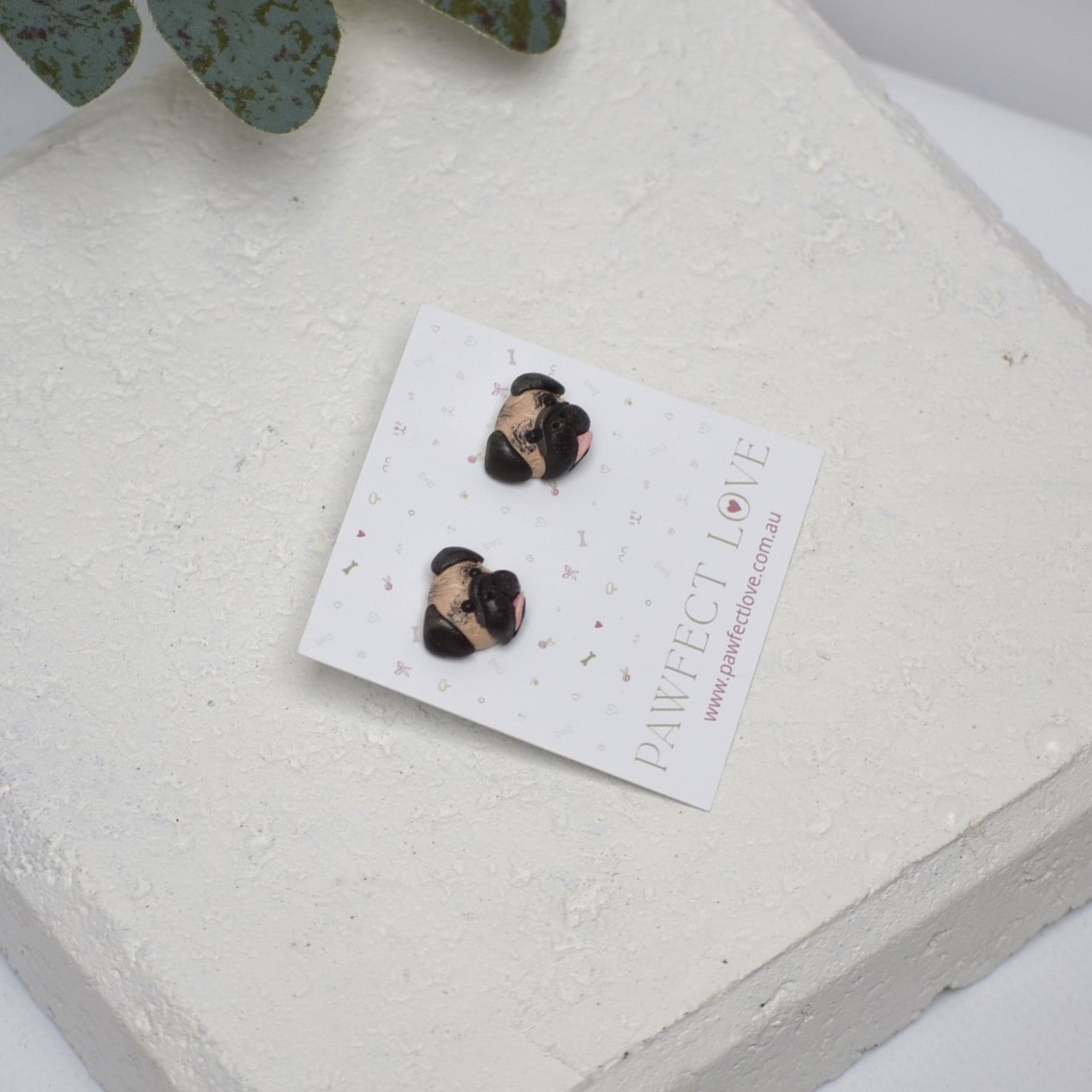 Handmade Pug stud earrings by Pawfect Love, positioned in front of white paver