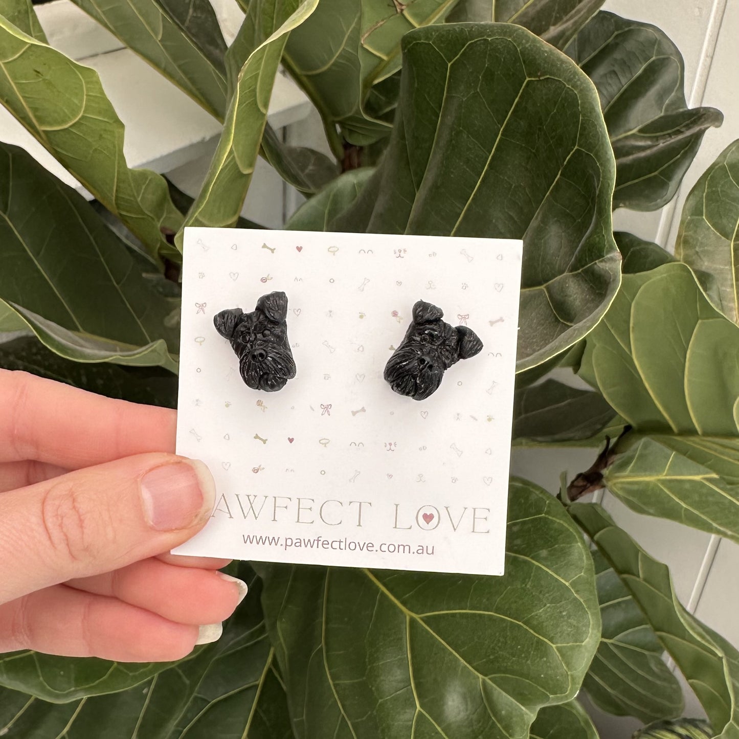 Black schnauzer dog-shaped earrings on a card held in front of green leaves