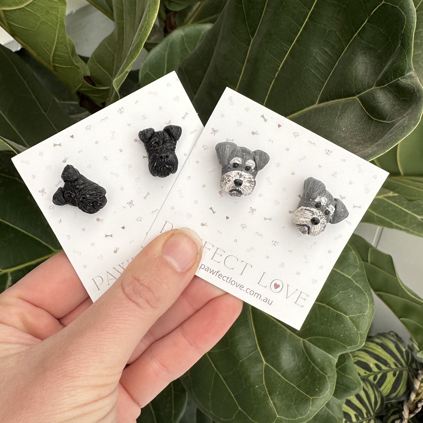 Pair of schnauzer dog-shaped earrings on display cards held by a hand with green leaves in the background.