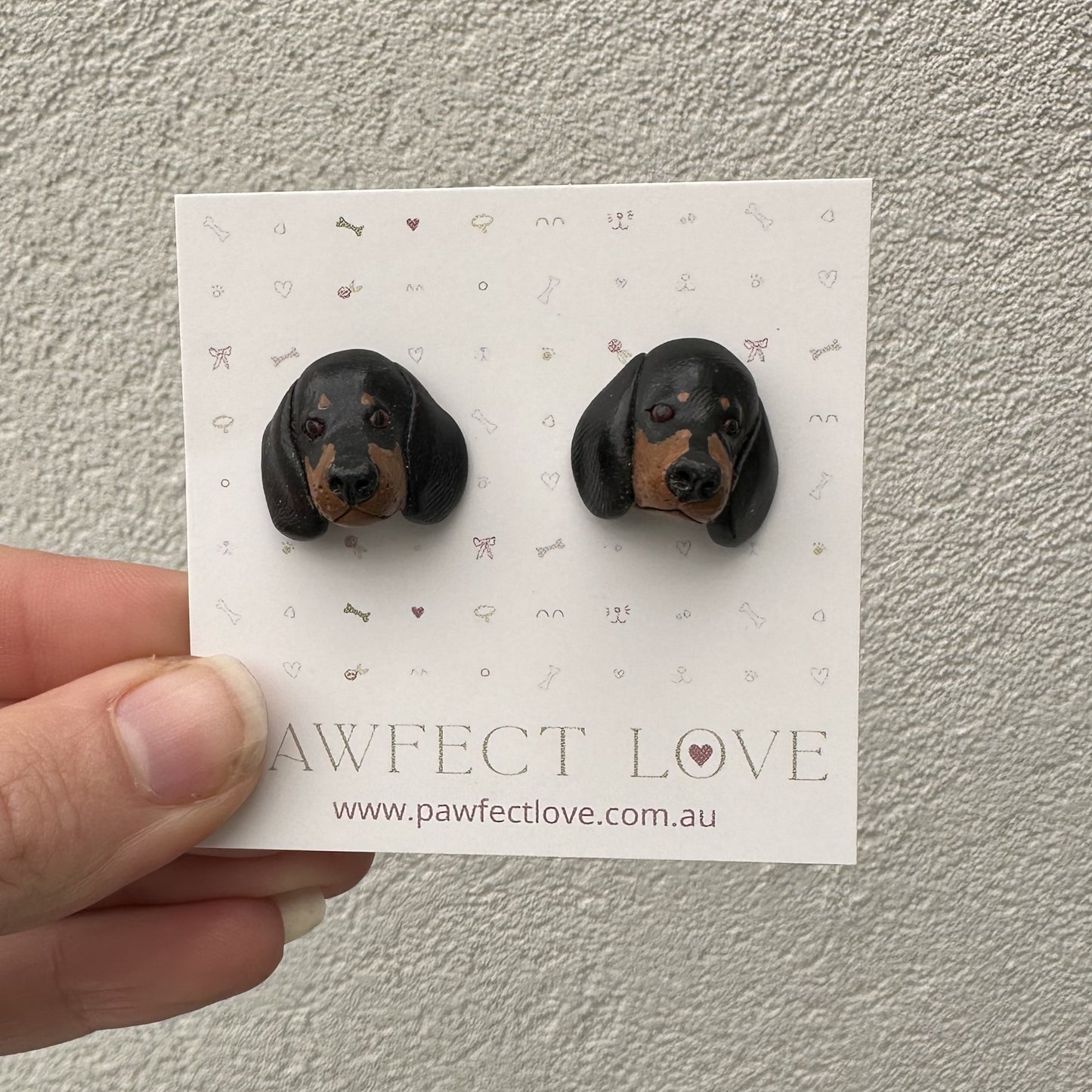 Black and brown dachshund dog-shaped earrings on a 'Pawfect Love' branded card held by a hand.