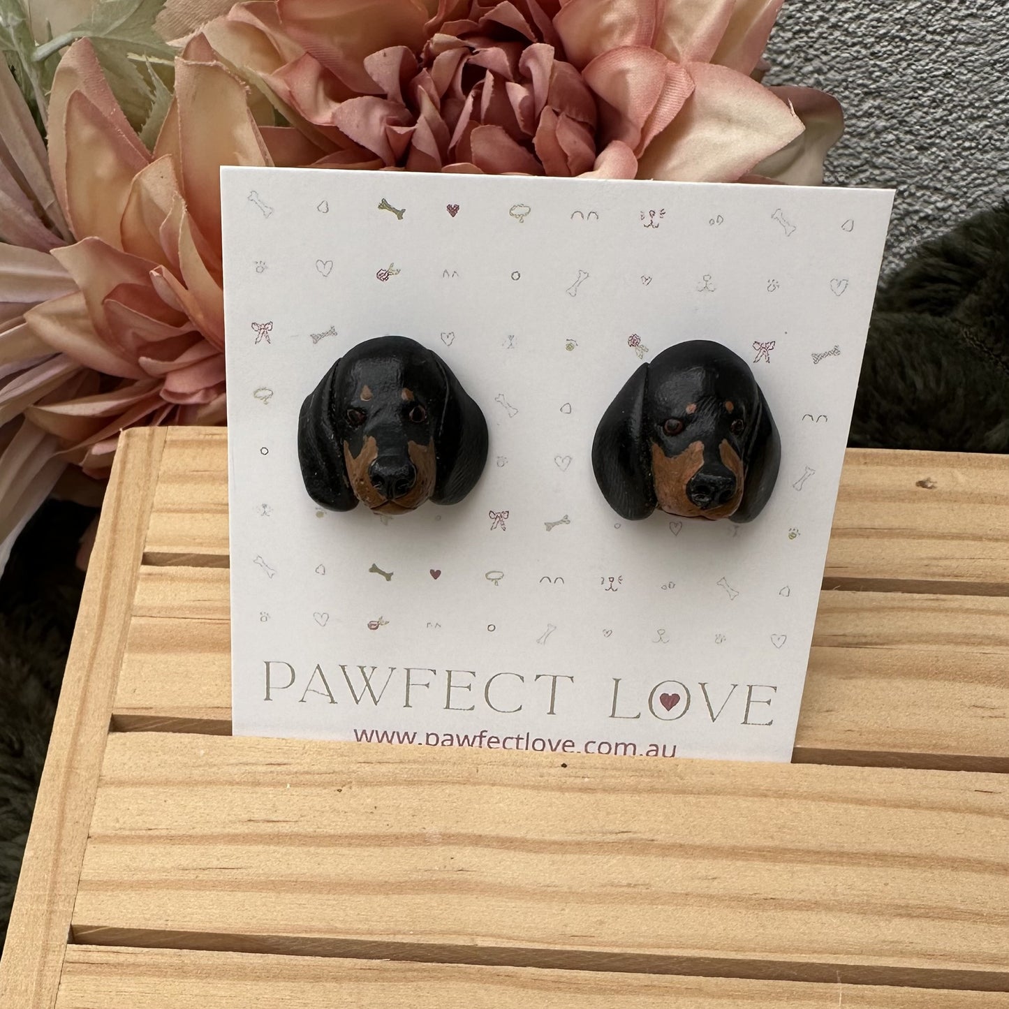 Black and brown dachshund dog-shaped earrings on a 'Pawfect Love' branded card with flowers in the background.