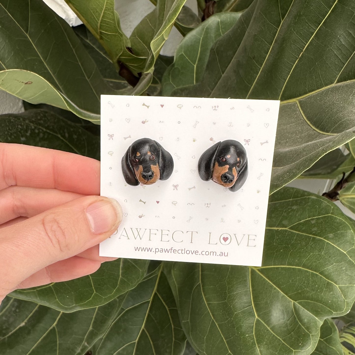 Pair of black and tan dachshund dog-shaped earrings on a 'Pawfect Love' branded card held over green leaves.