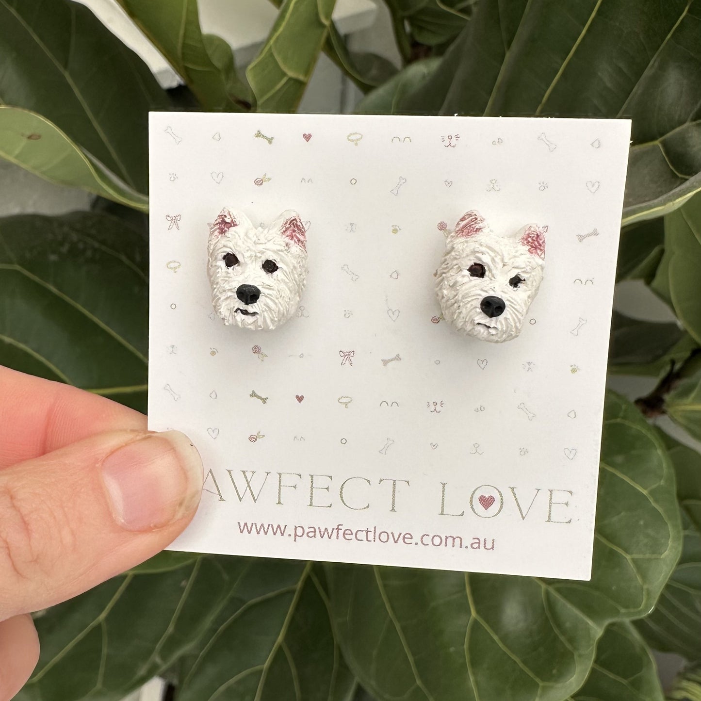 White Westie dog-shaped earrings on a 'Pawfect Love' branded card held against a green leafy background.