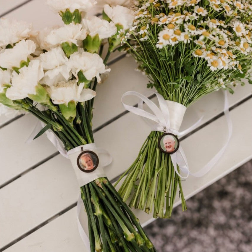 Two timber memorial photo charms of father and grandmother wrapped around bridal bouquet, pictured on a white bench.