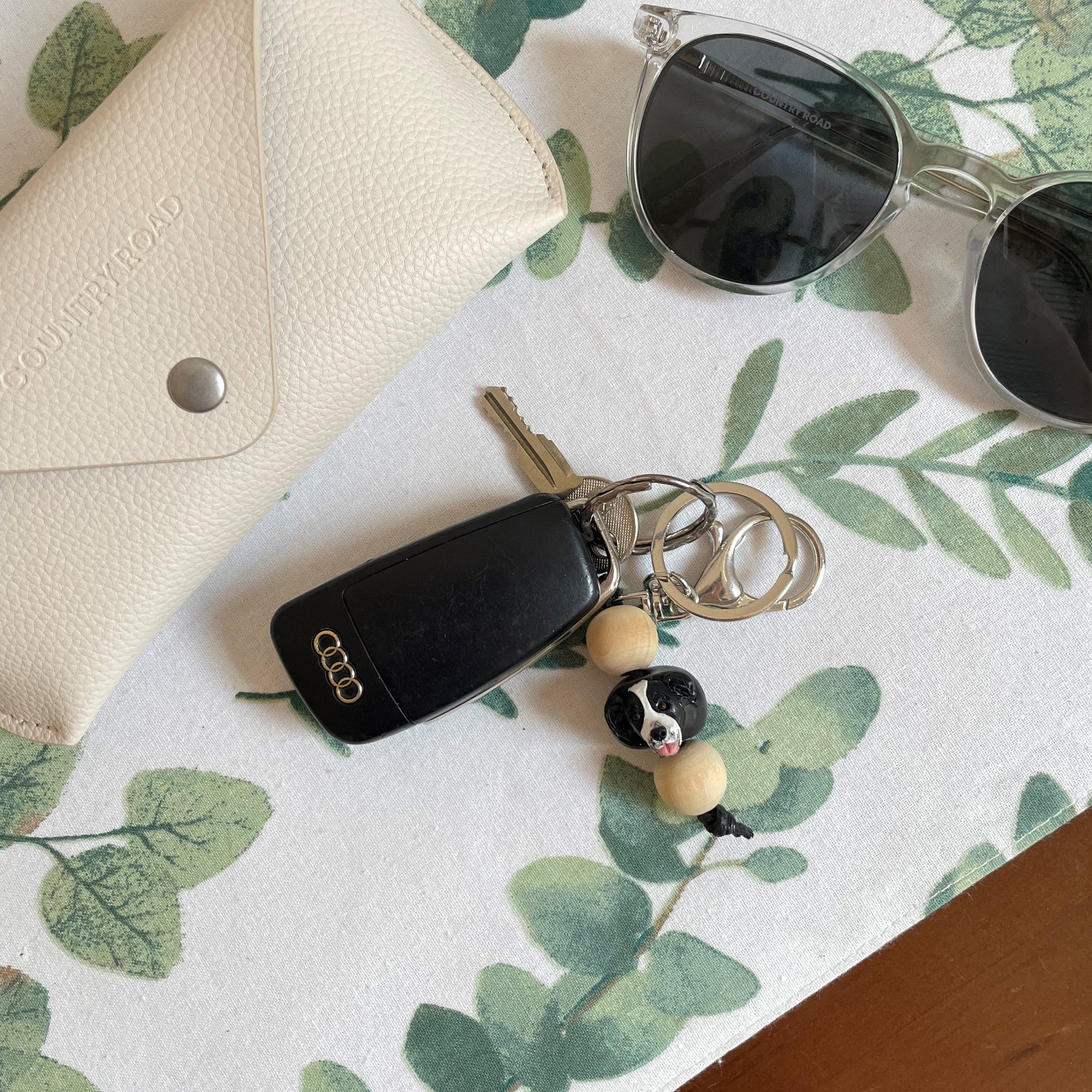 Handmade timber and polymer clay border collie dog keychain on a white table cloth beside sunglasses and case