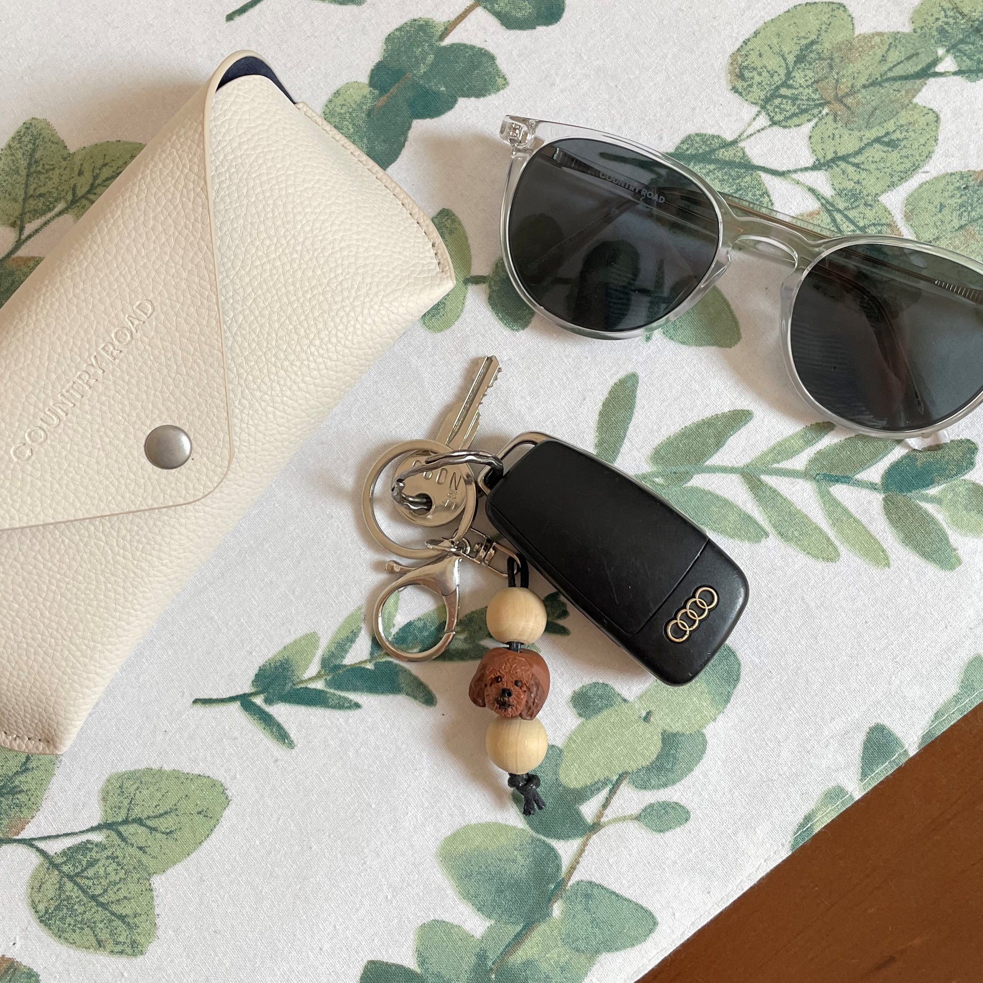 Handmade timber and polymer clay cavoodle dog keychain laying beside sunglasses and case