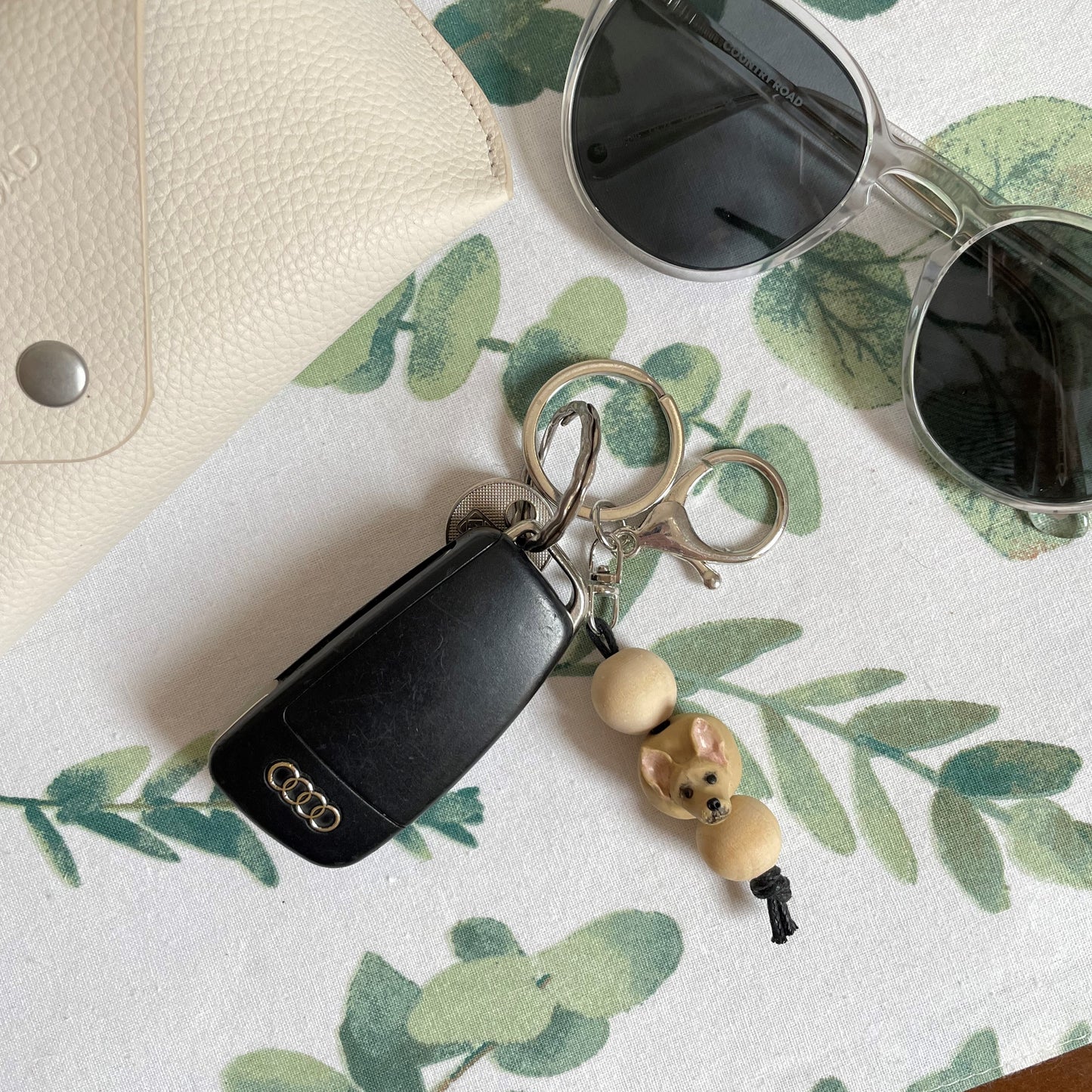 Handmade timber and polymer clay chihuahua dog keychain laying on white tablecloth beside sunglasses