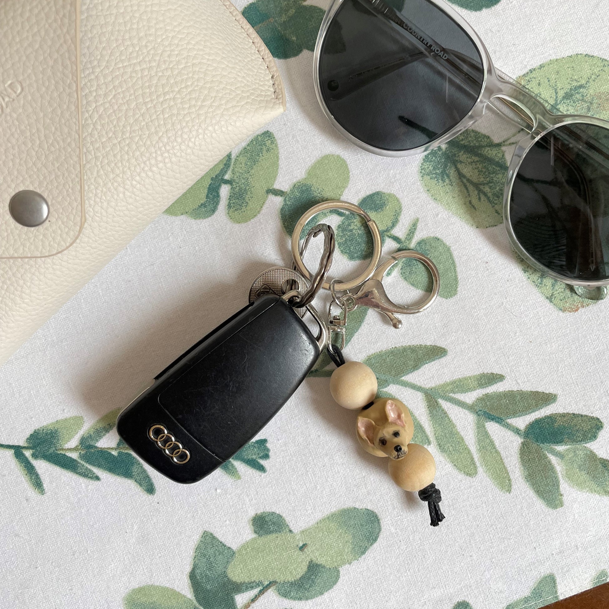 Handmade timber and polymer clay chihuahua dog keychain laying on white tablecloth beside sunglasses