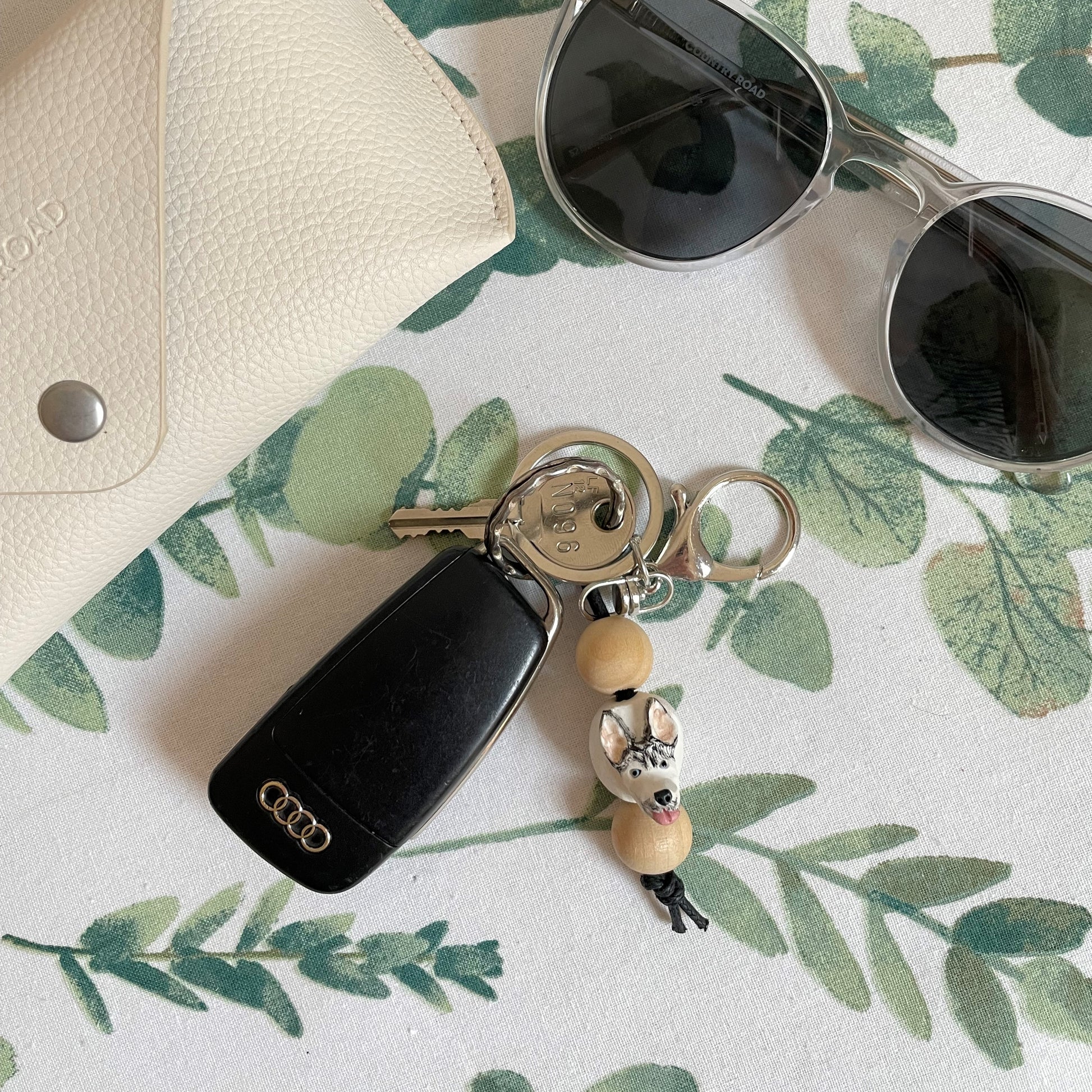 Handmade timber and polymer clay husky dog keychain on white floral tablecloth beside sunglasses