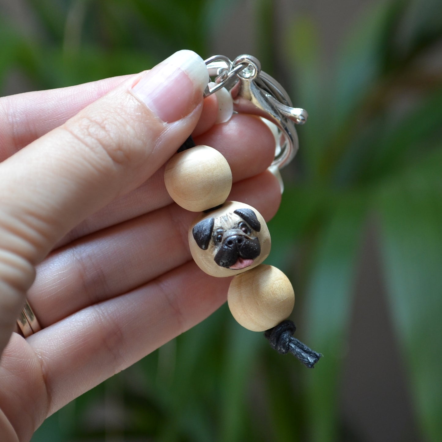 Handmade timber and polymer clay pug dog keychain being held in front of green palm plant background