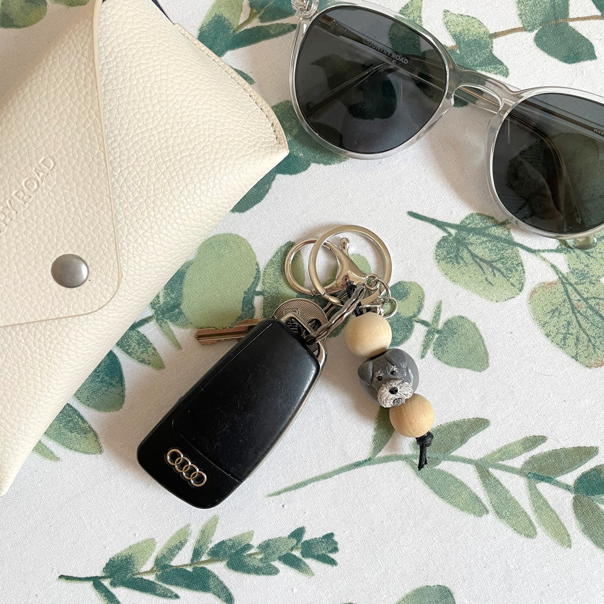 Handmade timber and polymer clay grey schnauzer dog keychain on white floral table runner beside sunglasses