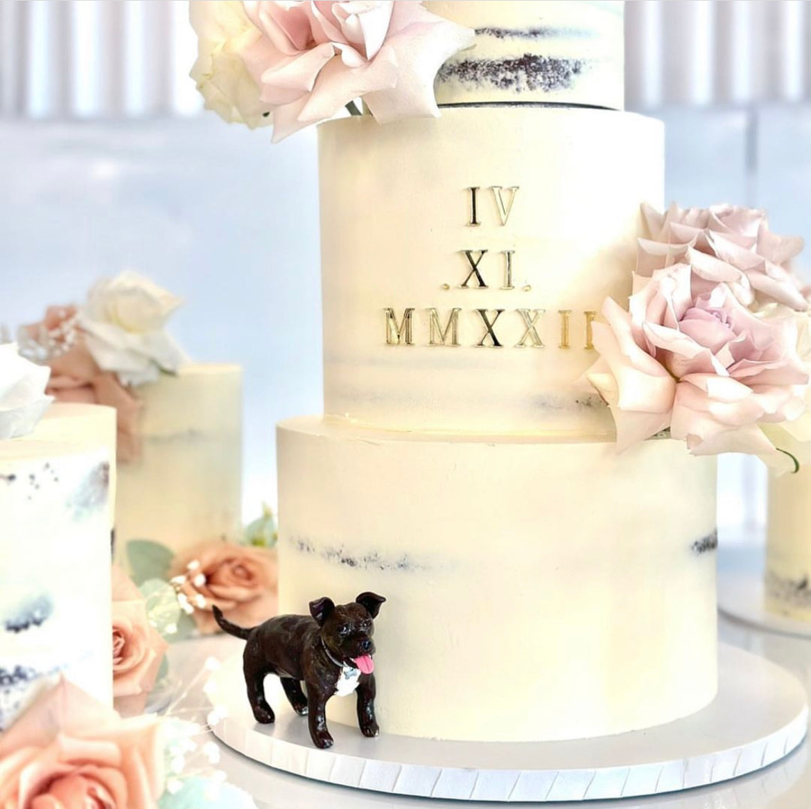 3 tier white white wedding cake with pink roses, surrounded by mini matching cakes with a handmade dog cake topper standing beside the main cake.