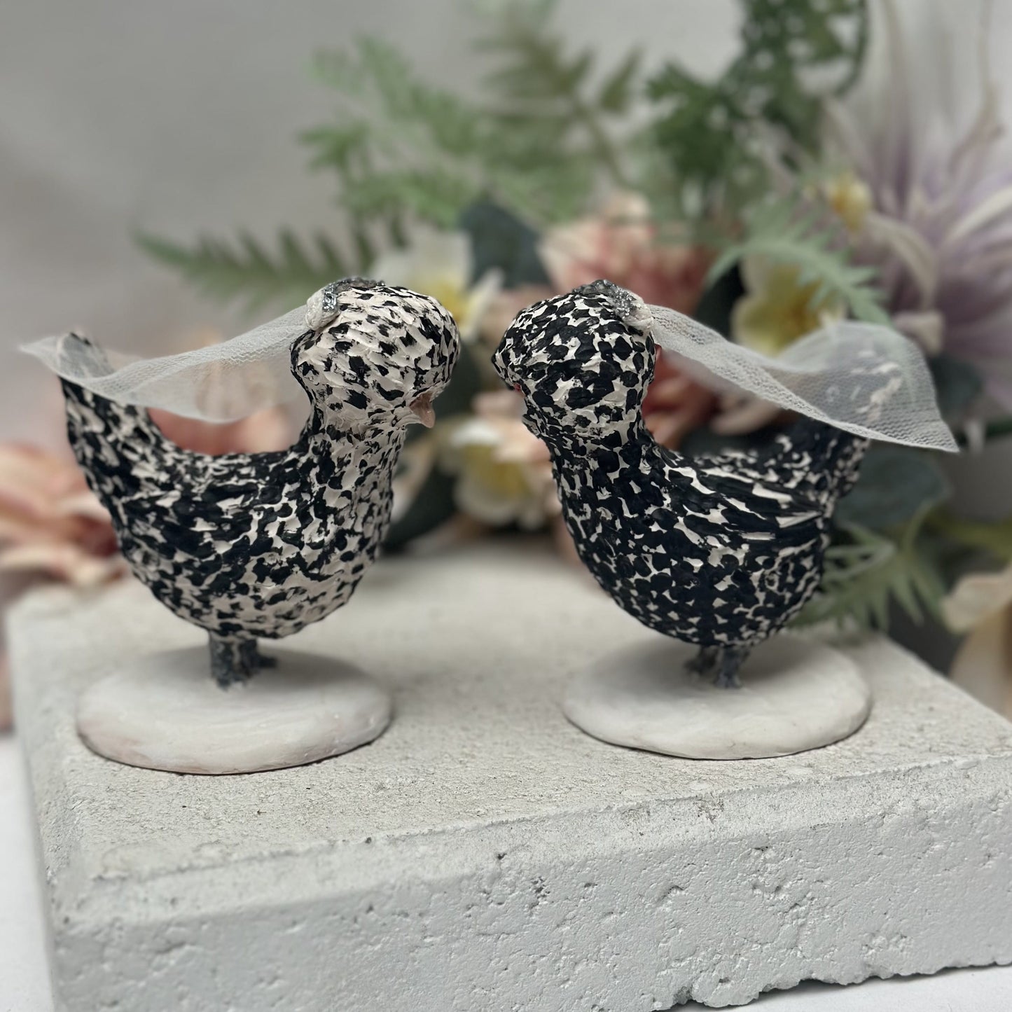 Two decorative bird figurines with lace on a stone surface with flowers in the background