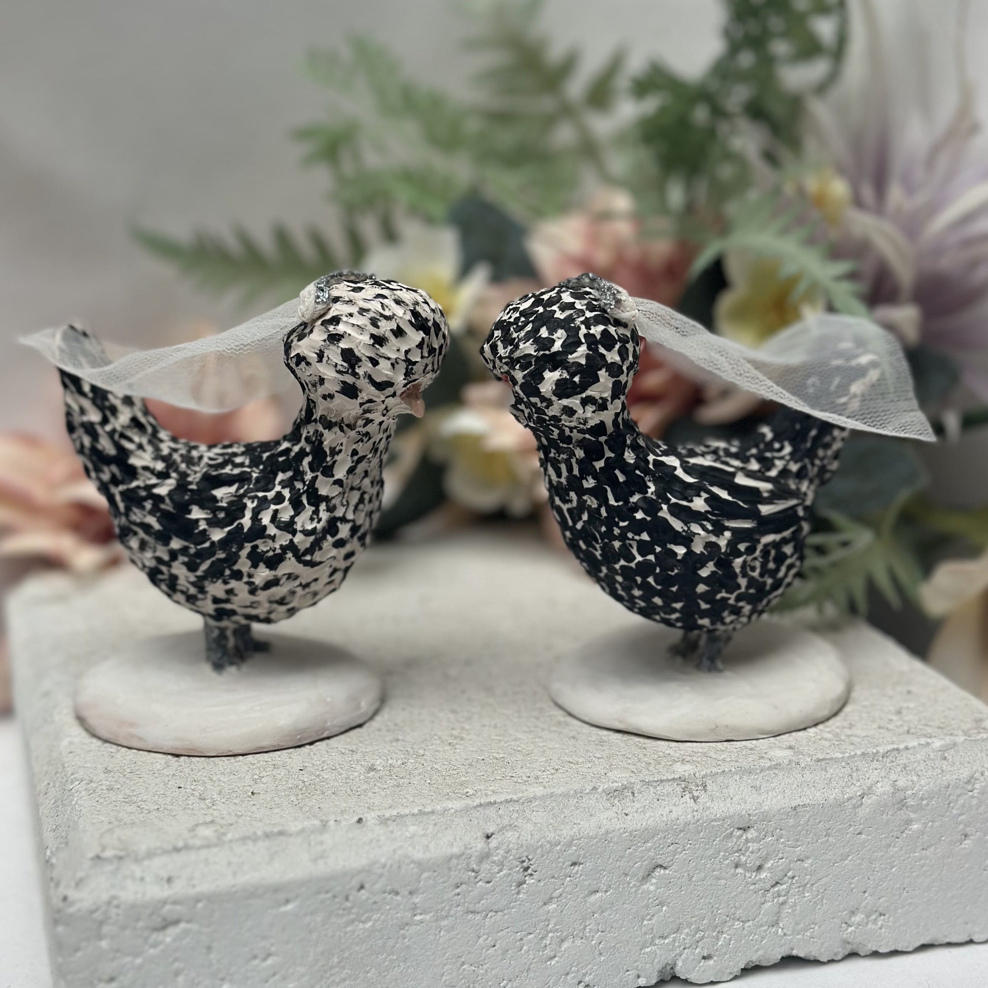 Two decorative bird figurines with lace on a stone surface with flowers in the background