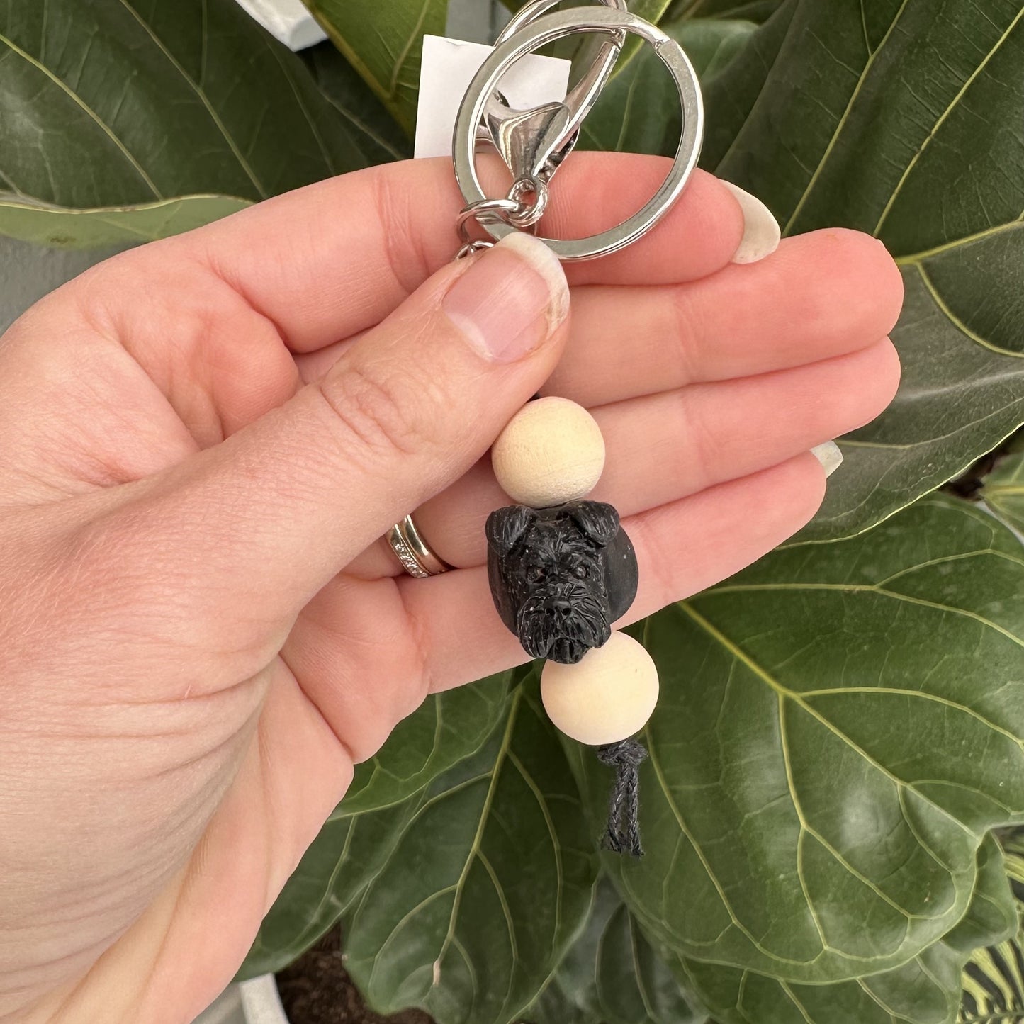 Hand holding a keychain with wooden beads and a black schnauzer face against a green leafy background