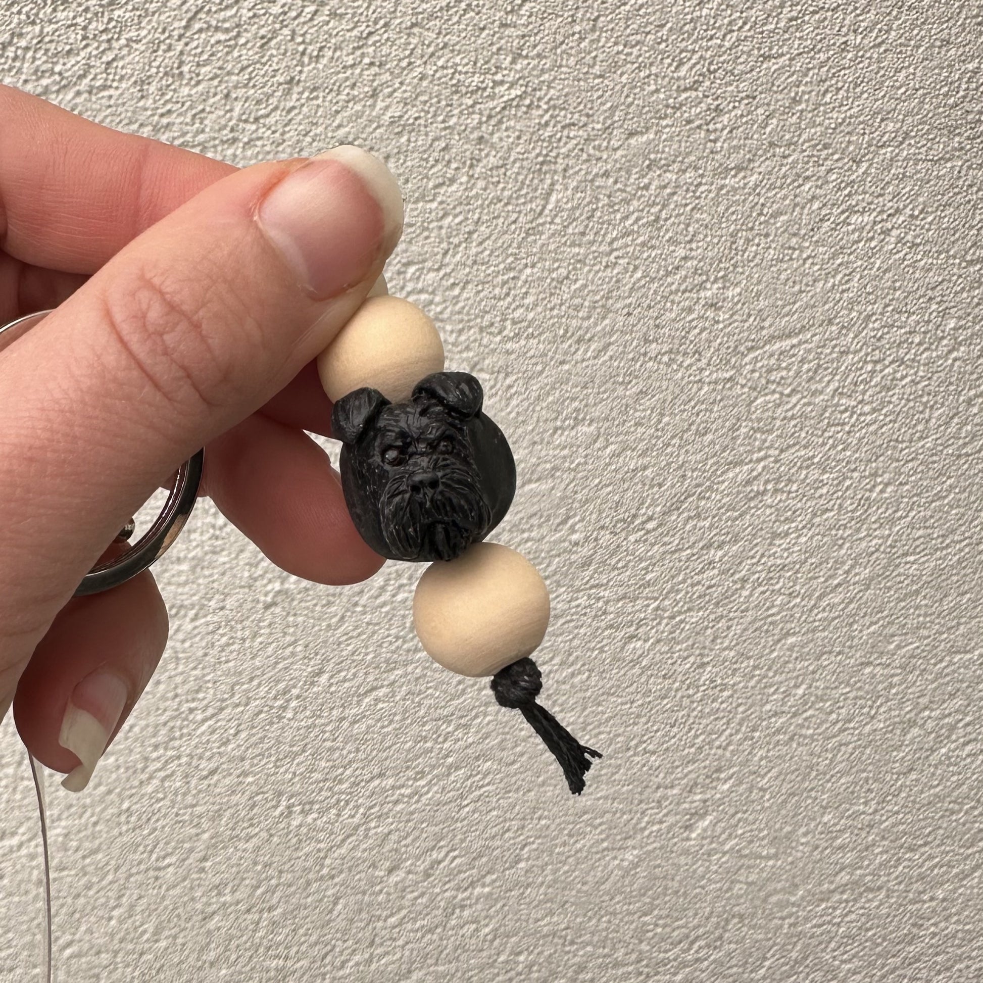 Hand holding a keychain with a black schnauzer dog figurine and two beige beads against a light background