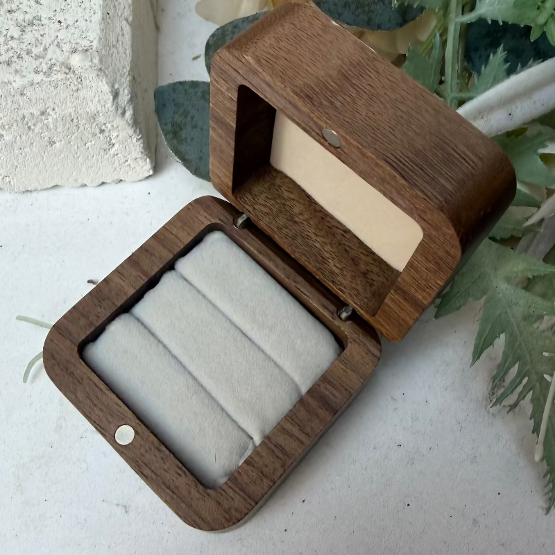 A small open wooden jewelry box with a dark brown wood-grain finish and a light beige interior, showing three padded cream fabric slots. The box is photographed on a light surface with decorative flowers and greenery in the background.