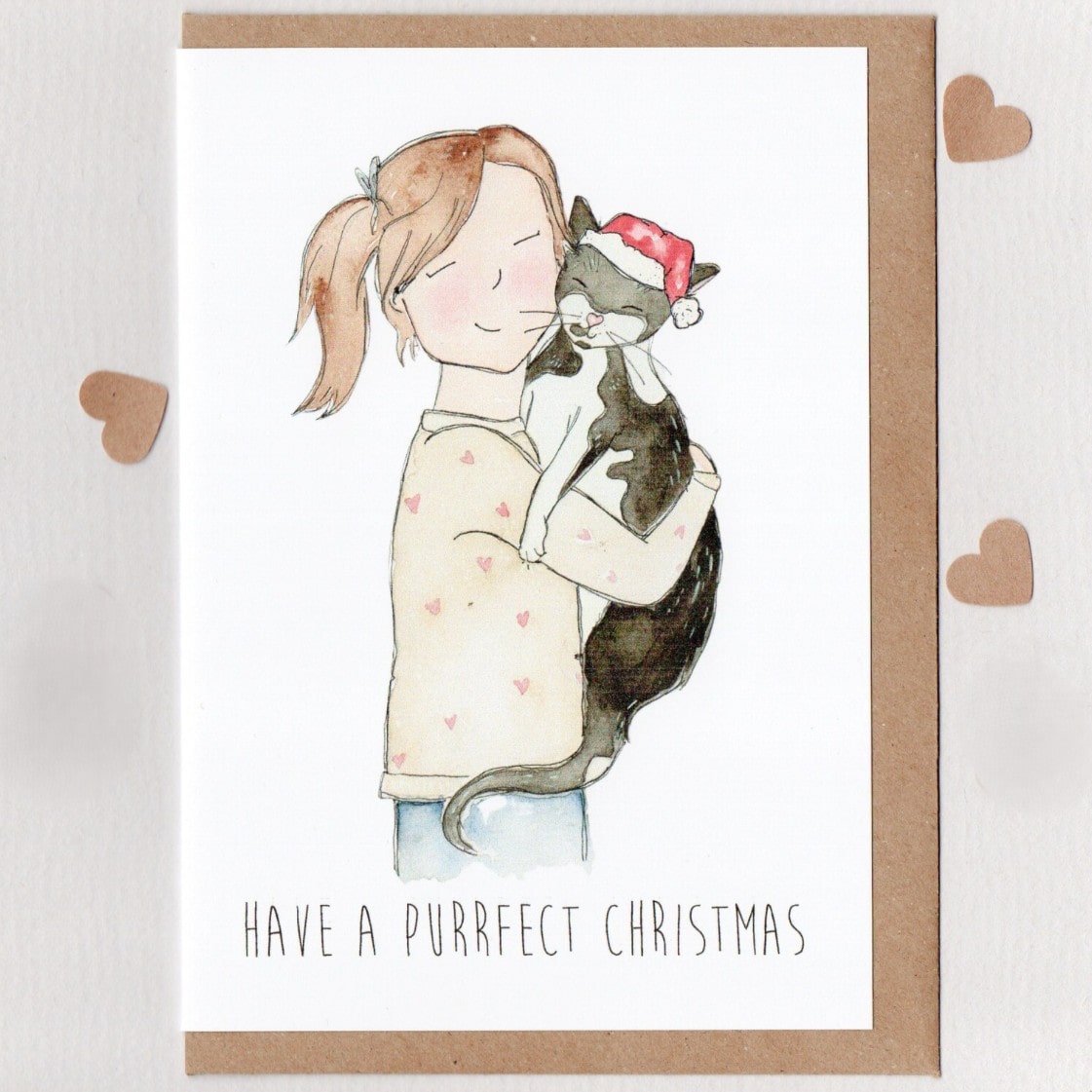 Card with illustration of a girl holding a cat wearing a Santa hat, text 'Have a purrfect Christmas', on a white background with brown border.