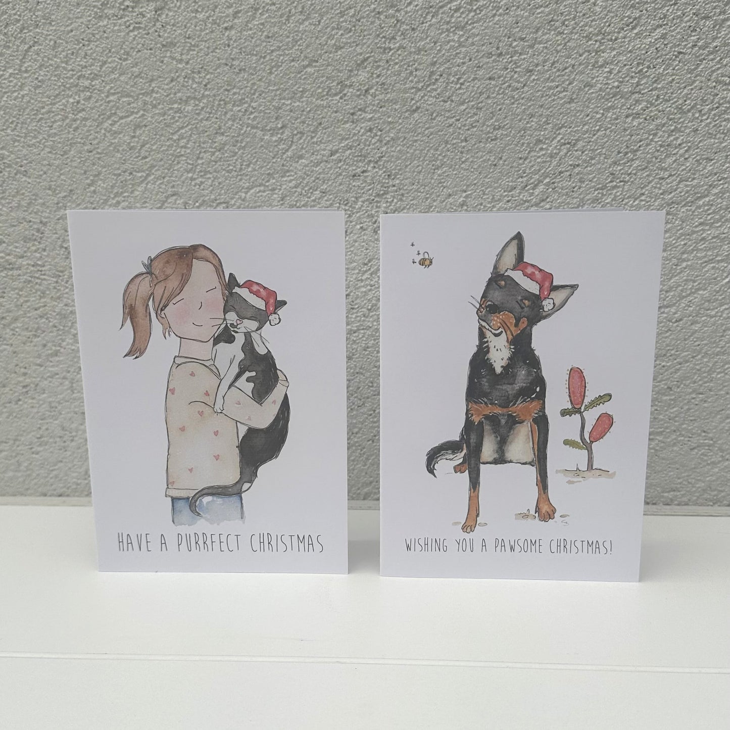Two Christmas-themed greeting cards with illustrations of a girl and a dog on a textured gray background.