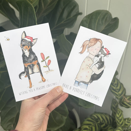 Two pet- themed Christmas cards held by a hand with plants in the background