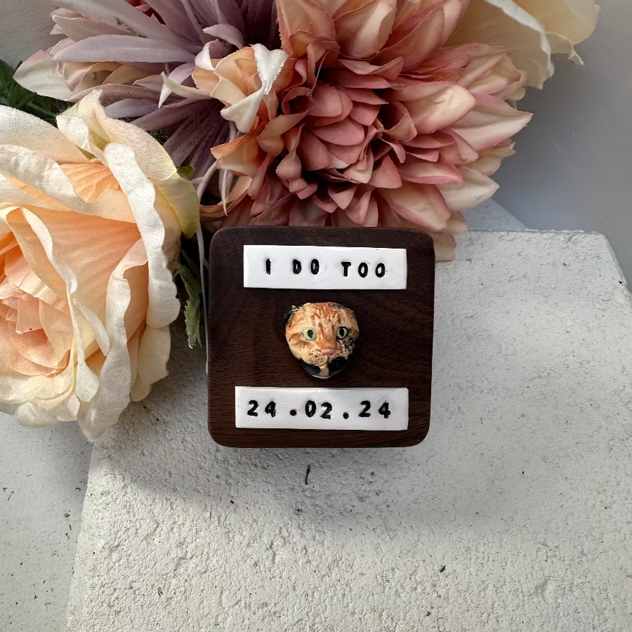 A small wooden keepsake box or plaque with a round glass vial containing a dried flower, displayed on a light gray surface with decorative artificial flowers behind it. The front has white labels reading 'I DO TOO' and the date '24.