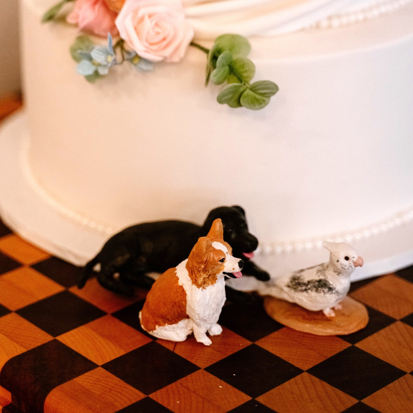 Decorative figurines of 2 dogs and a bird wedding cake topper on a checkered surface with a cake in the background.