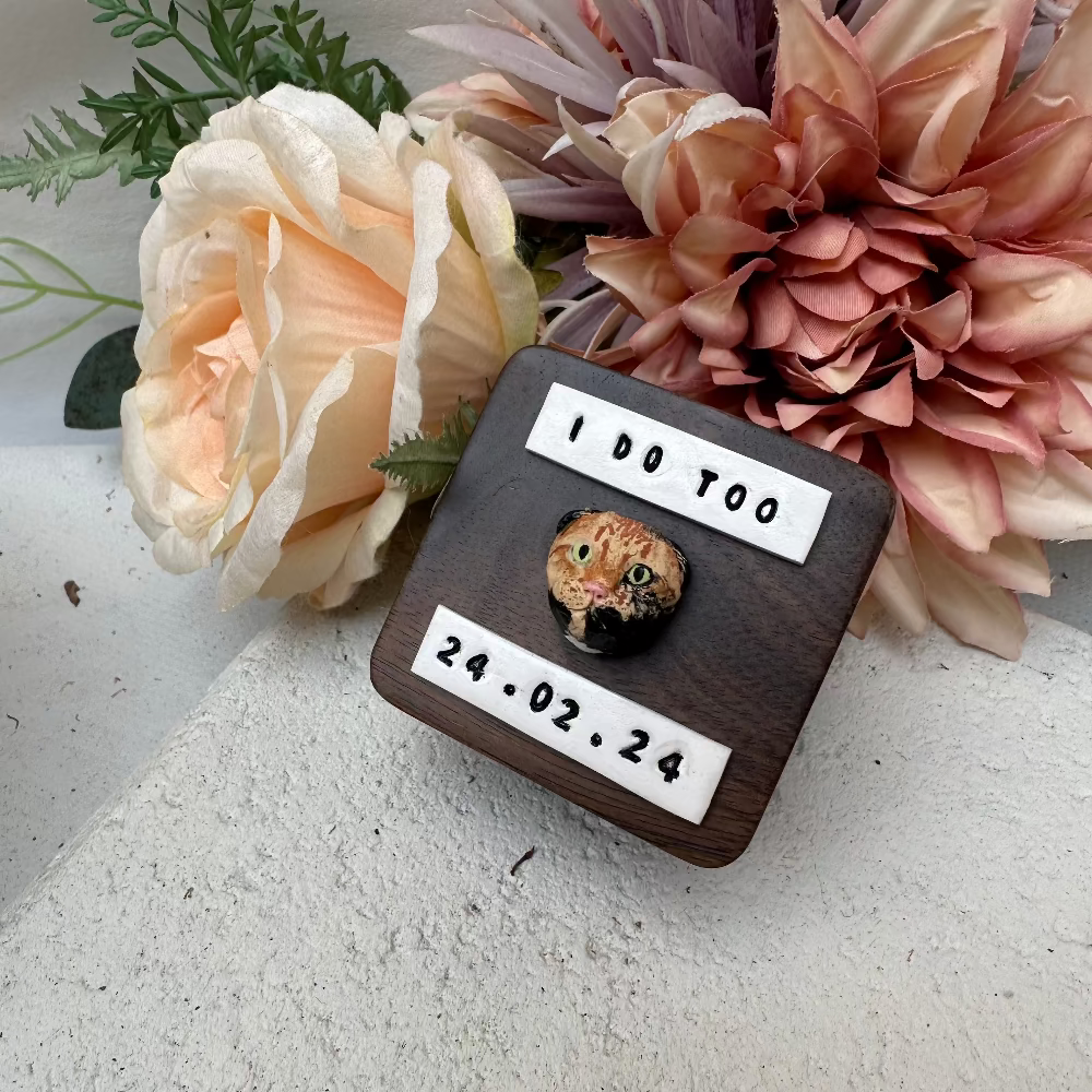 A bespoke double ring box with a custom pet face design, made of wood, with a faux fur lining. The box is inscribed with a date and time, and displays a ring inside it, placed on a floral arrangement.