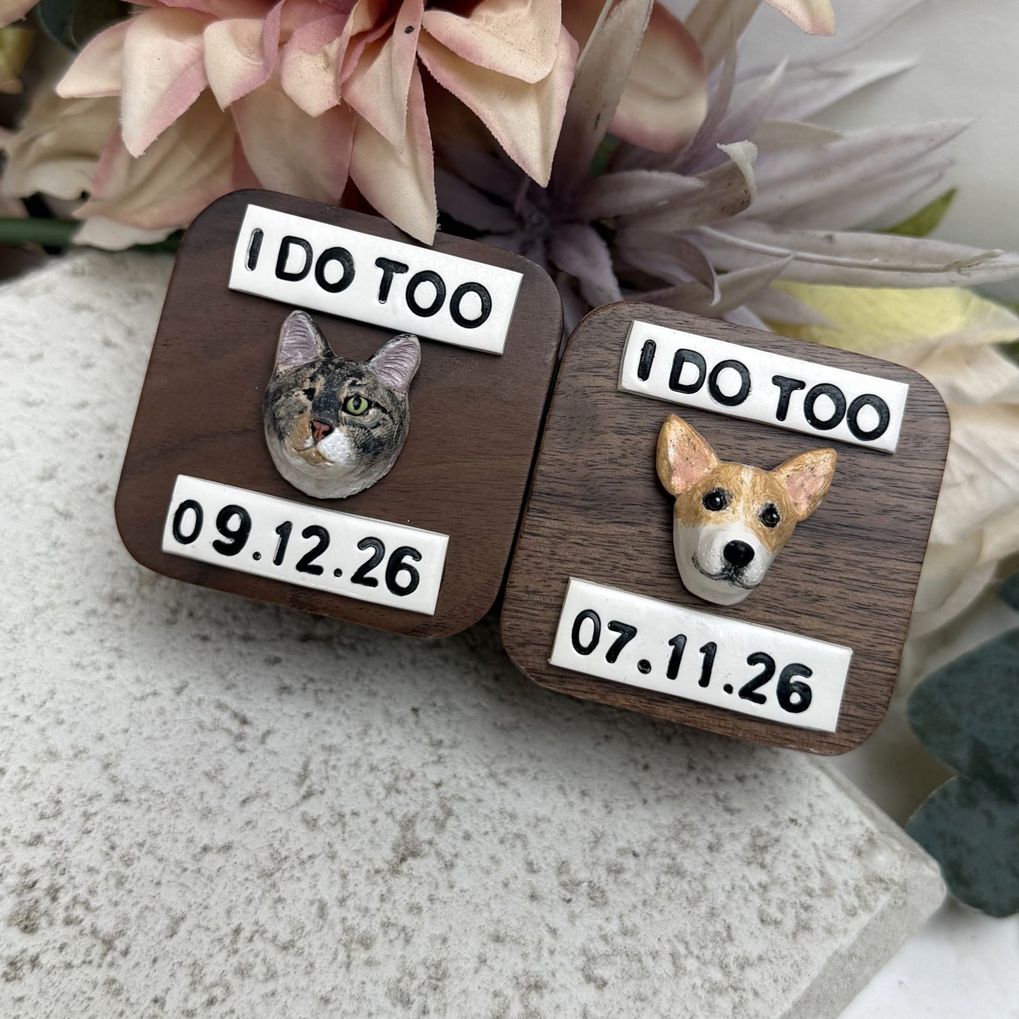 Two square wooden ring boxes with rounded corners displayed on a stone surface with dried flowers in the background. One box features a cat portrait and the other features a dog portrait, both with white labels reading 'I DO TOO' and engraved dates.