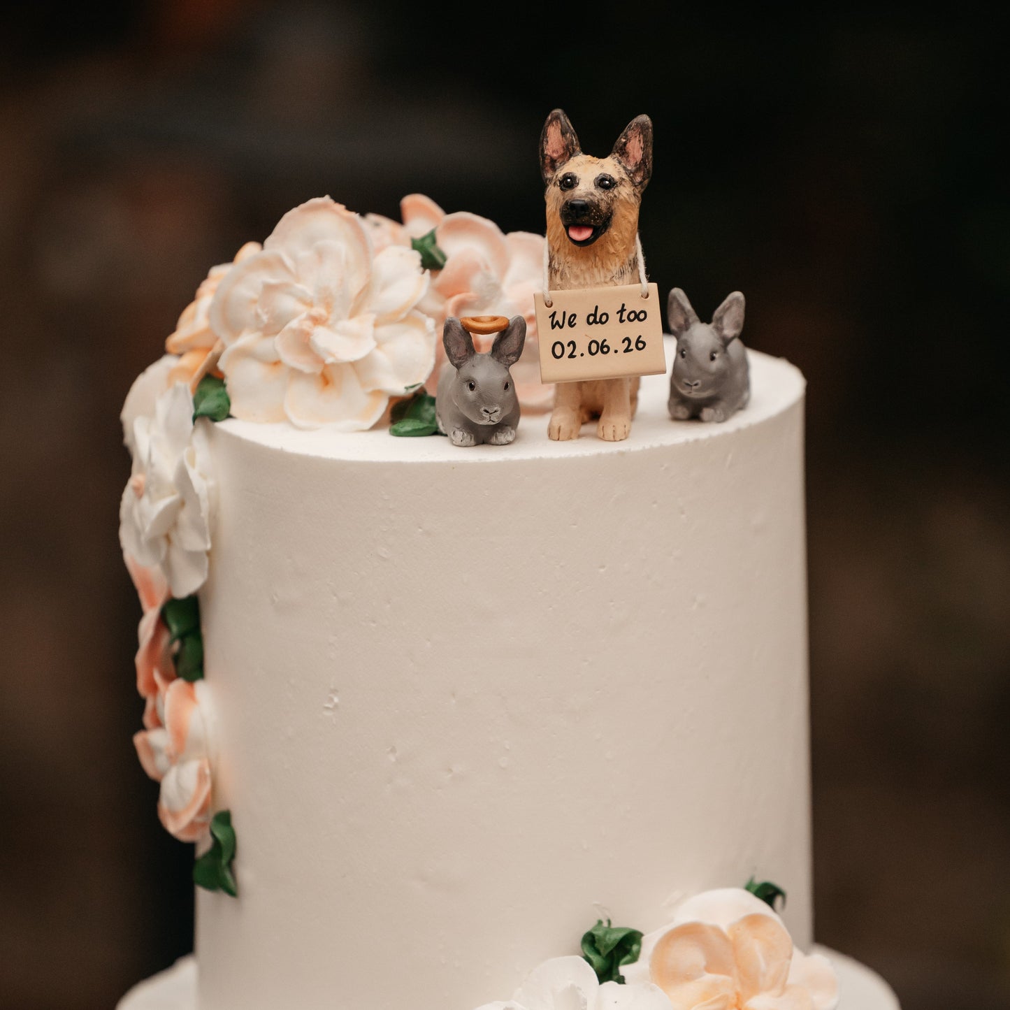 A close-up of a white two-tier cake decorated with peach and white flowers, green leaves, and small figurines on top, including a dog holding a small sign that reads 'We do too 02.06.