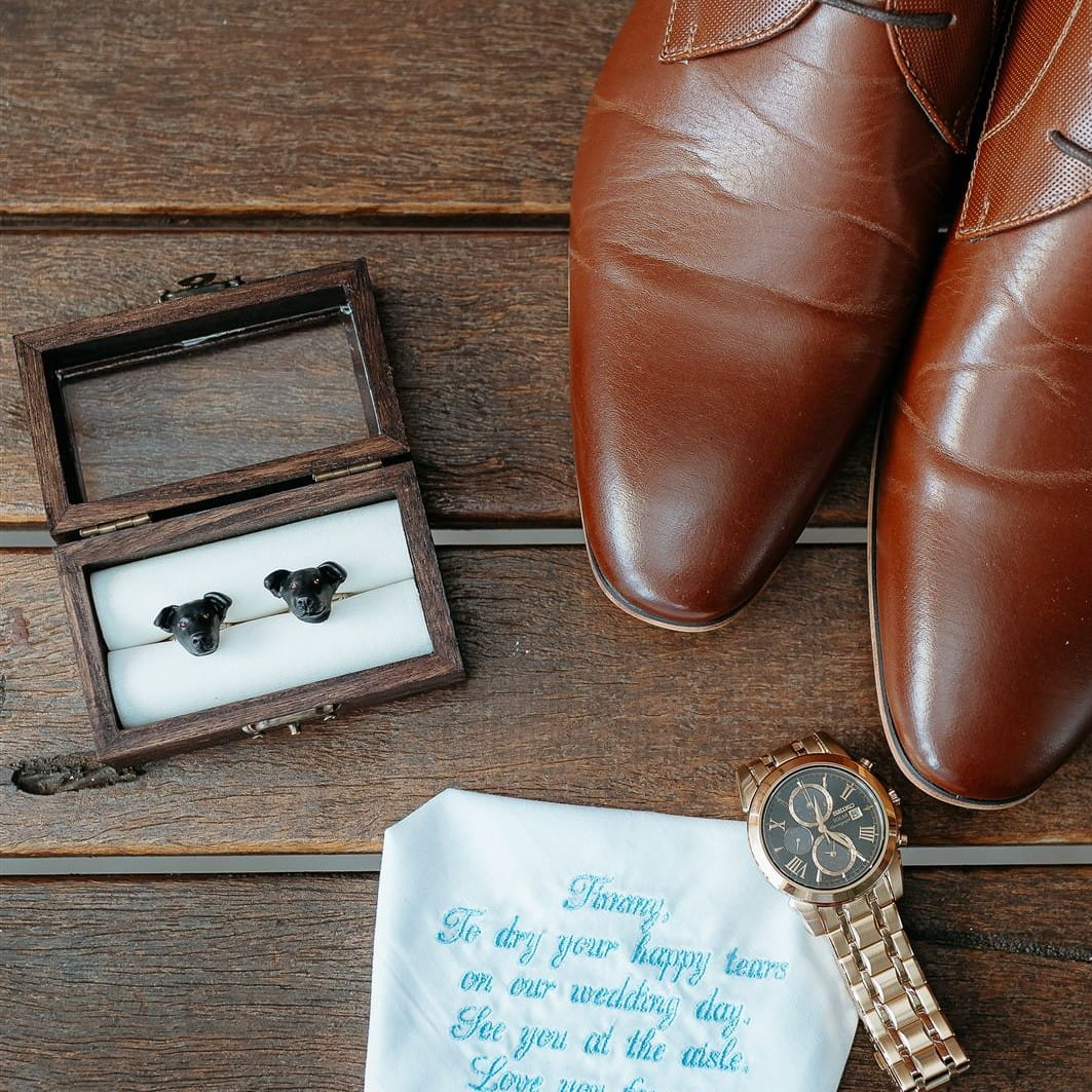 Brown leather shoes with a wooden box containing black Pawfect Love custom dog cufflinks, a watch, and a white handkerchief on a wooden surface.