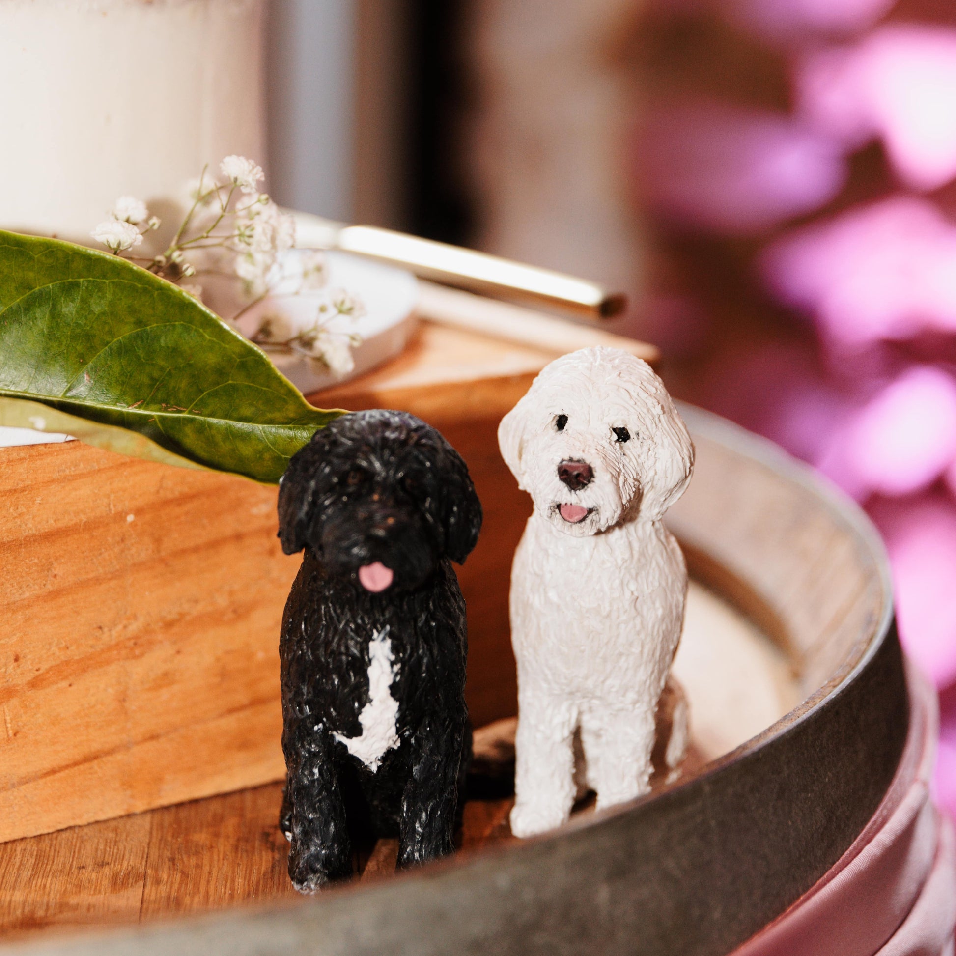 Two small handmade groodle and labradoodle cake topper dog figurines, one black and one white, sitting on a wooden surface with a blurred floral background.