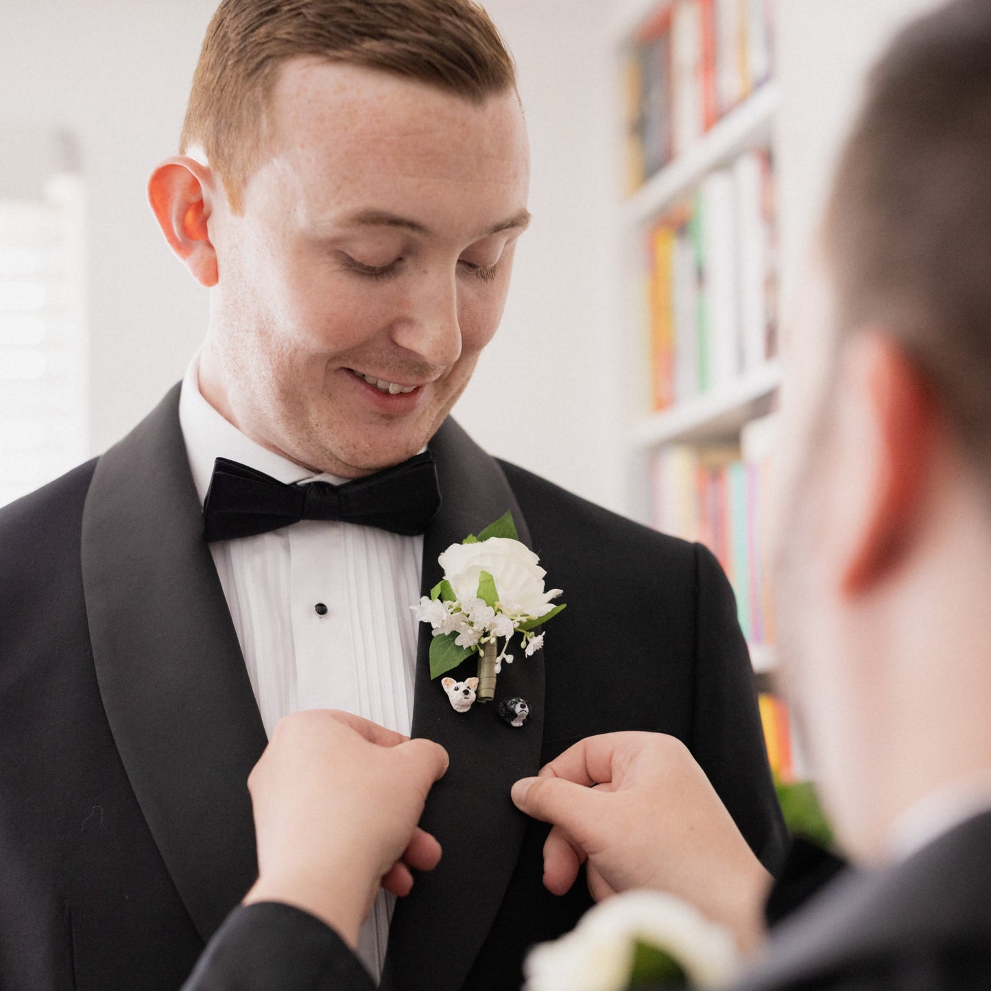 Man in tuxedo with floral boutonniere being adjusted by another person in a library setting, featuring 2 custom dog lapel pins