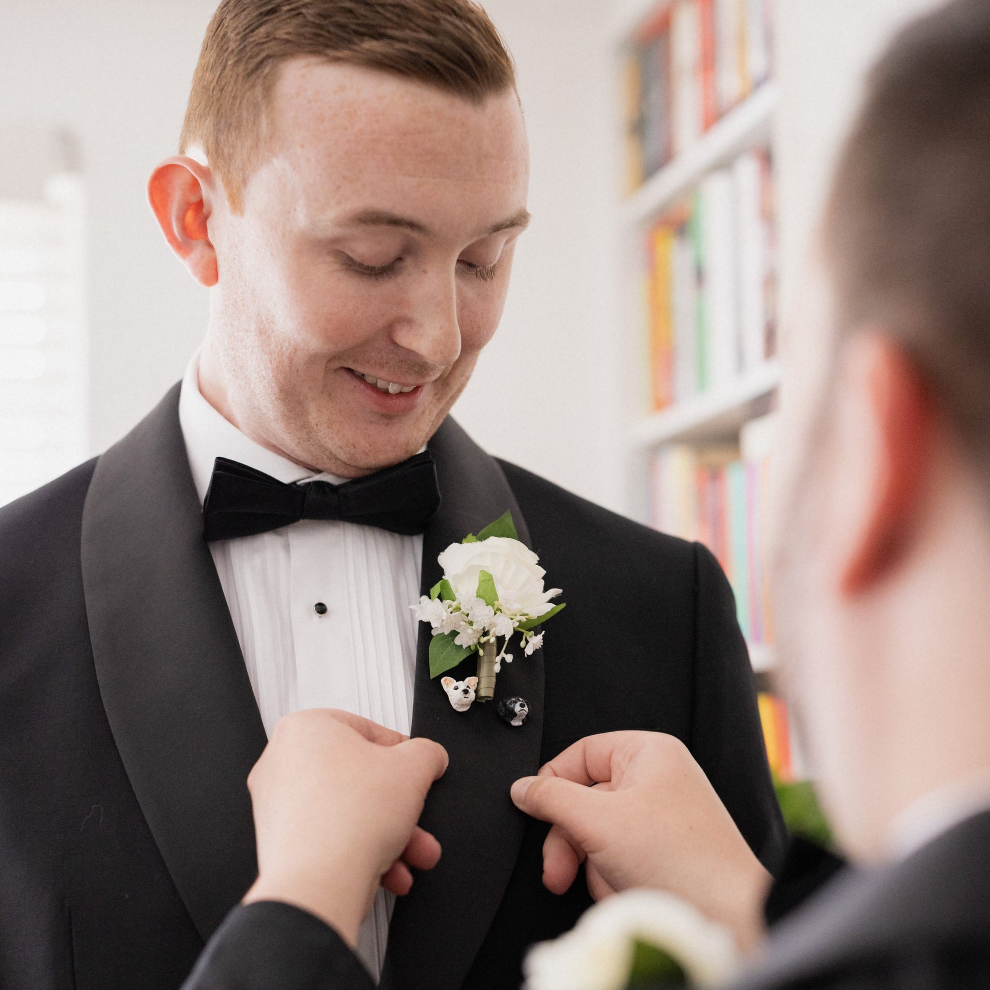 Man in tuxedo with floral boutonniere being adjusted by another person in a library setting, featuring 2 custom dog lapel pins