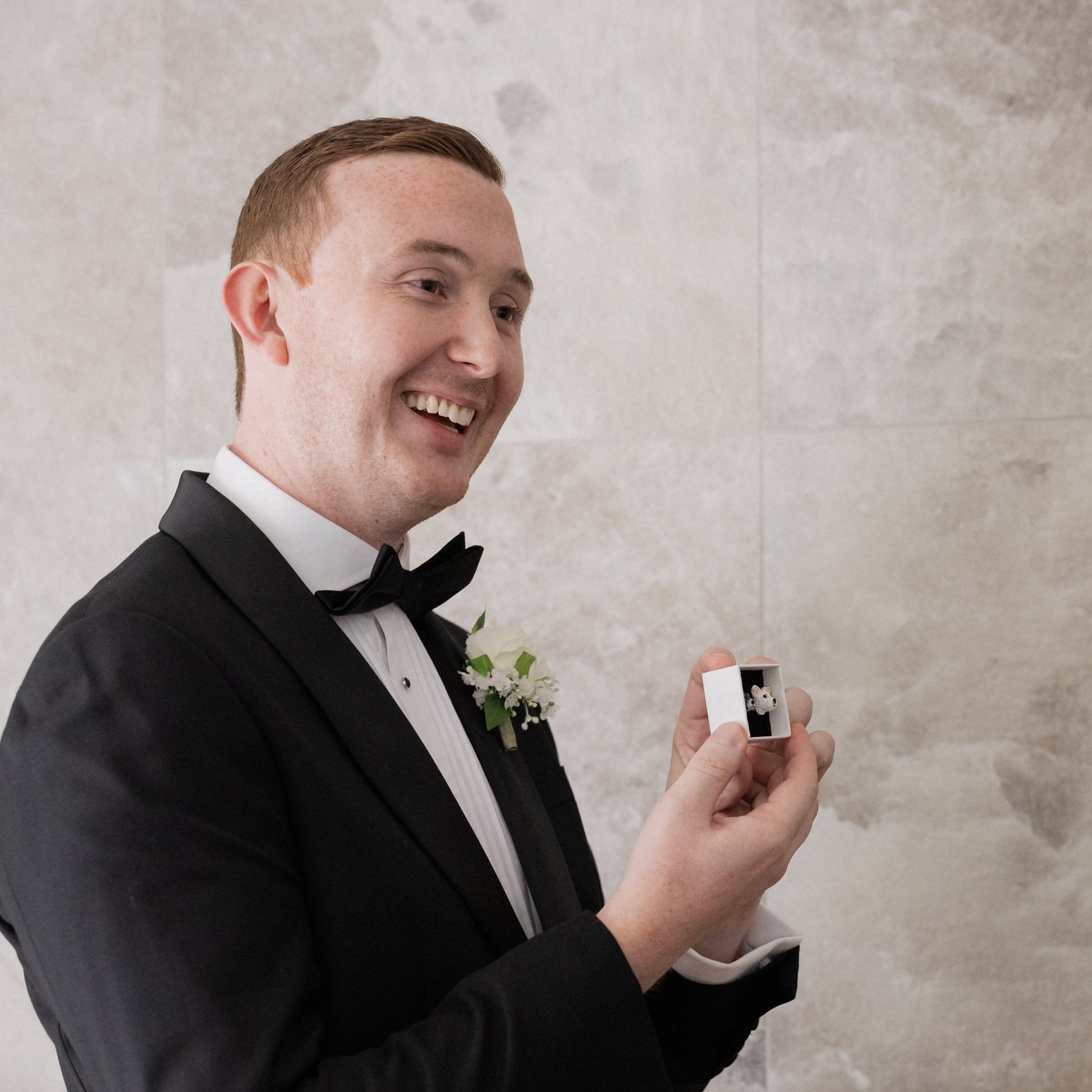 Man in a tuxedo holding a small box with custom dog lapel pins, smiling against a neutral background