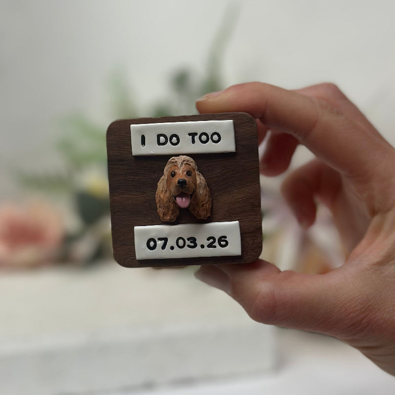 Hand holding a wooden timber wedding ring box with a dog's photo and text, against a blurred background.