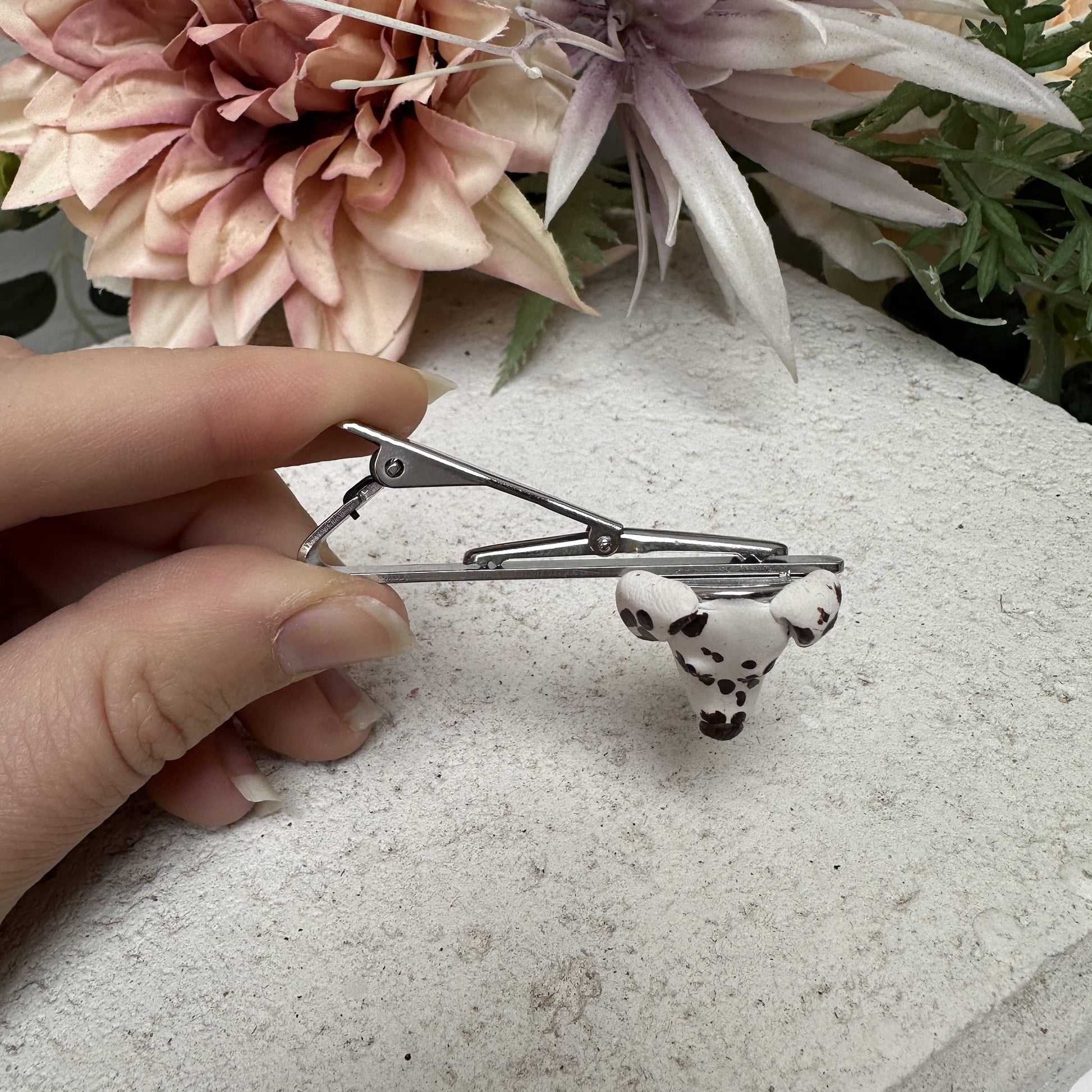 Hand holding a custom dog tie clip with a small dalmatian dog charm against a floral background.