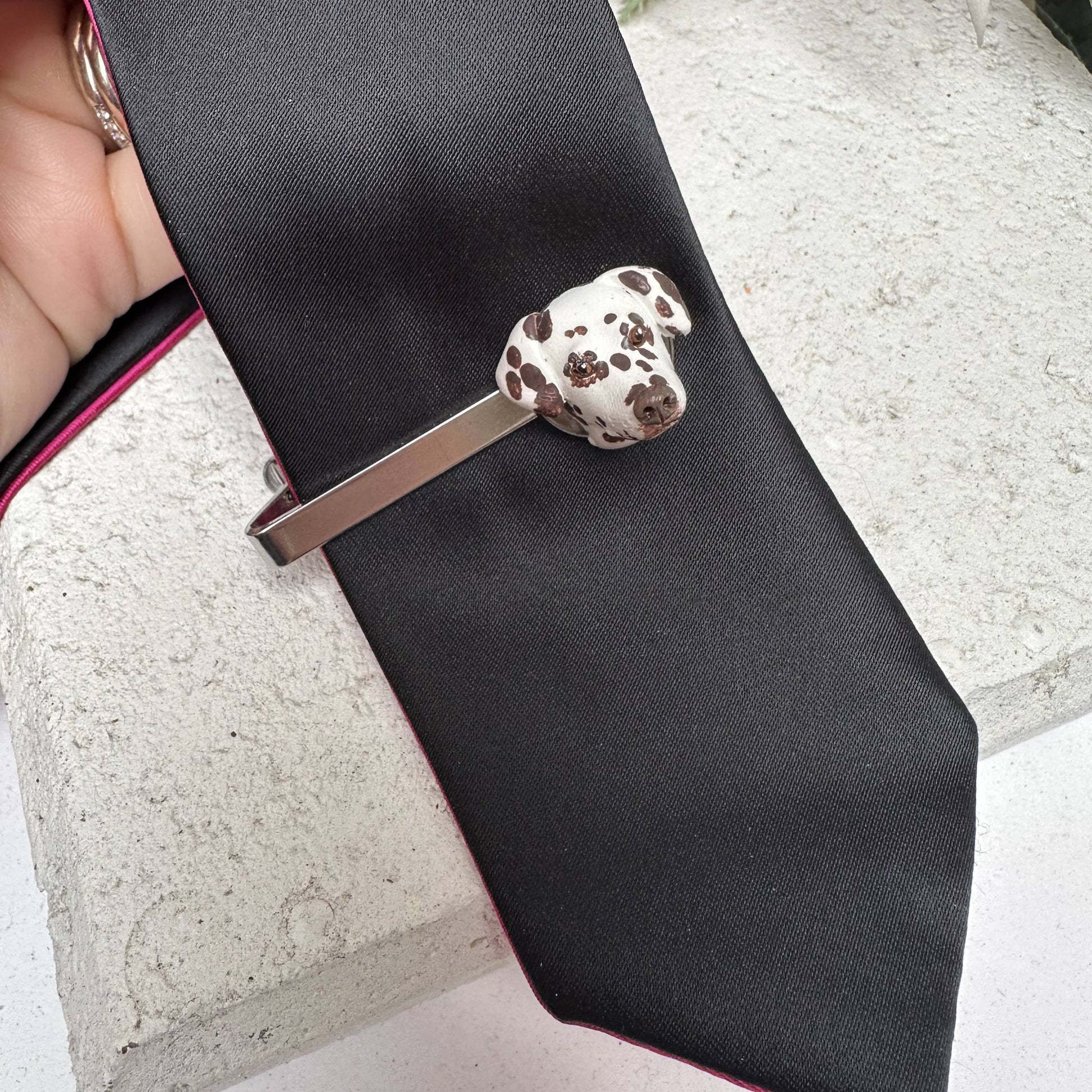 Tie clip with a handmade custom pet dalmatian face on a black tie against a stone surface