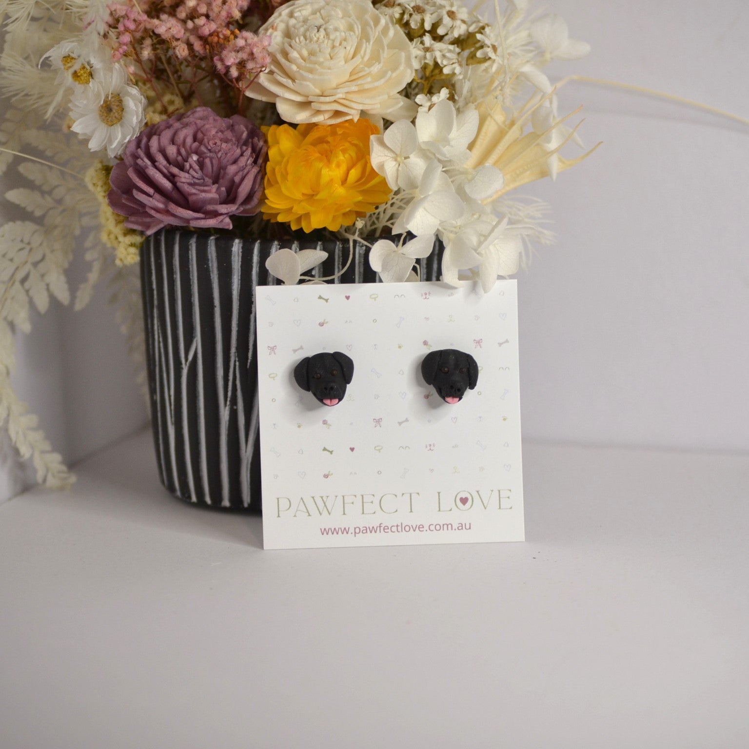 Handmade black labrador stud earrings by Pawfect Love, positioned in front of dried flower arrangement