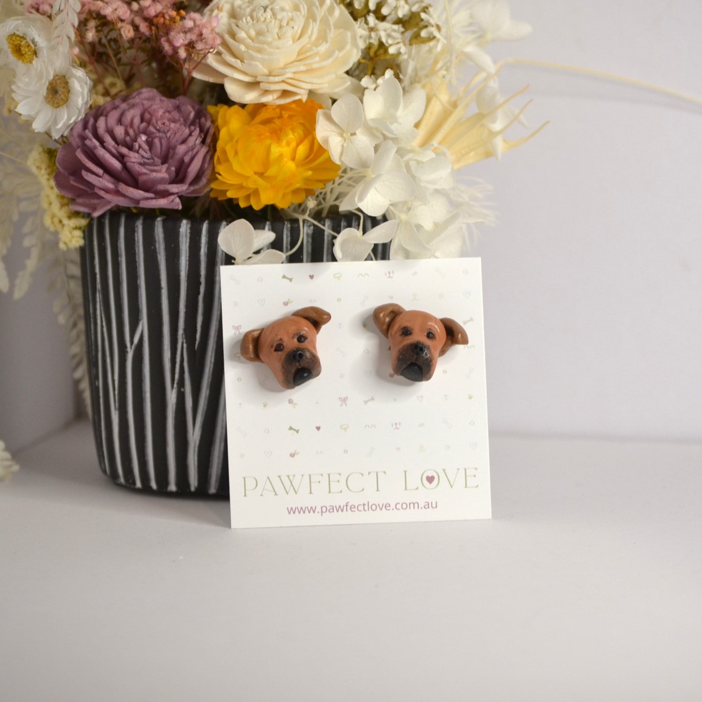 Handmade staffie stud earrings by Pawfect Love, positioned in front of dried flower arrangement