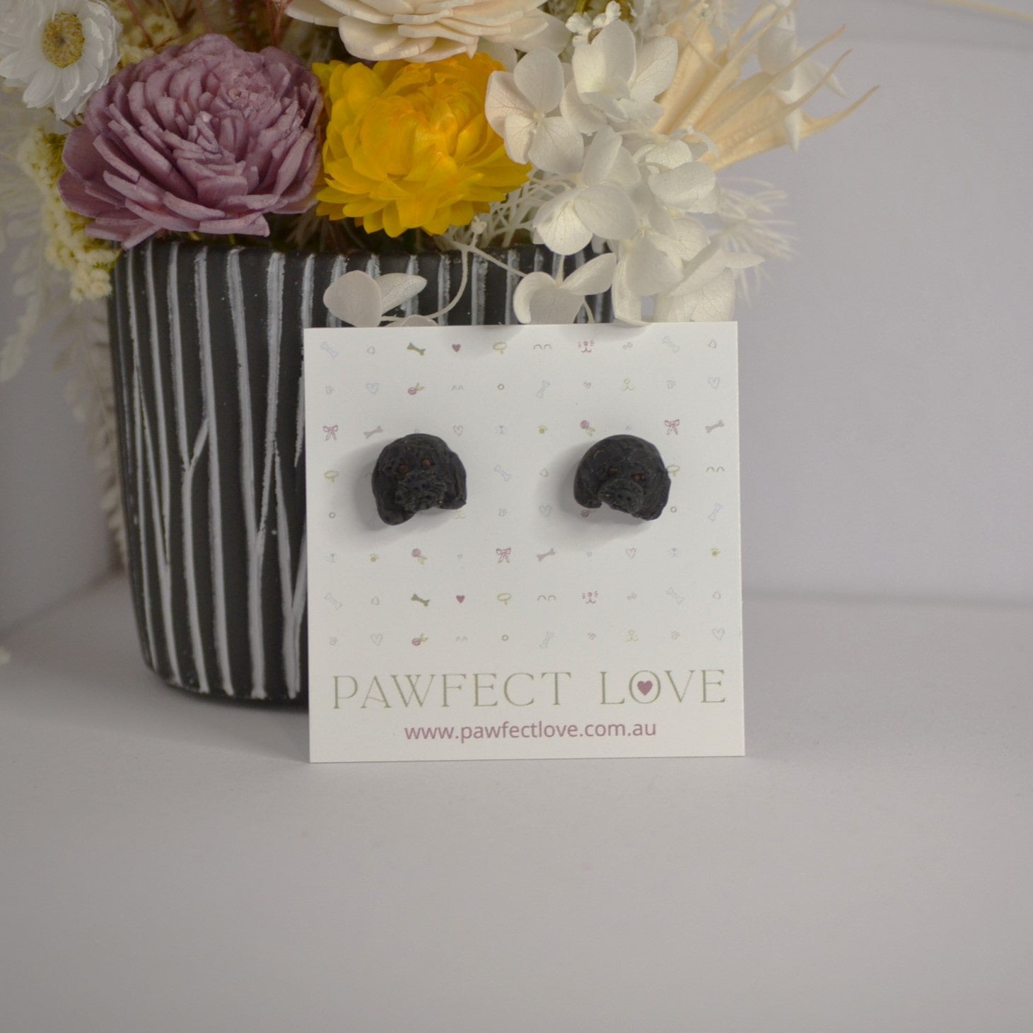 Handmade black poodle stud earrings by Pawfect Love, positioned in front of dried flower arrangement