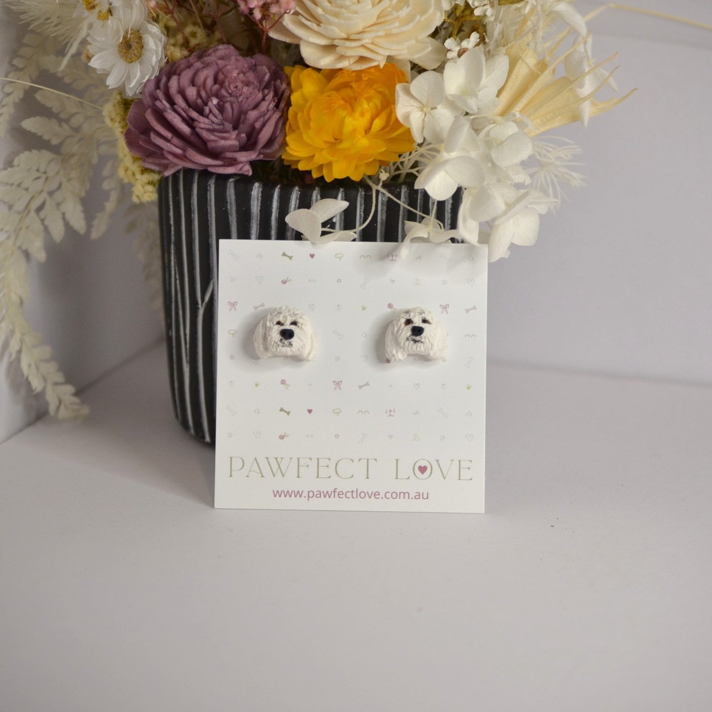 Handmade white poodle stud earrings by Pawfect Love, positioned in front of dried flower arrangement