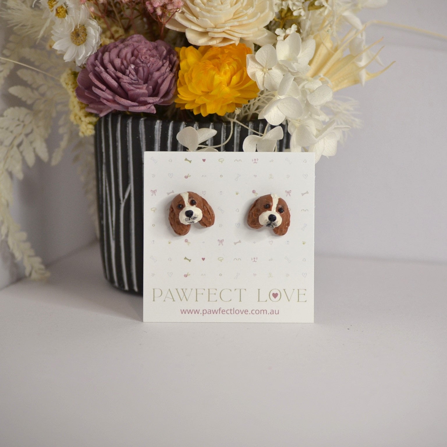 Handmade cavalier king charles spaniel stud earrings by Pawfect Love, positioned in front of dried flower arrangement