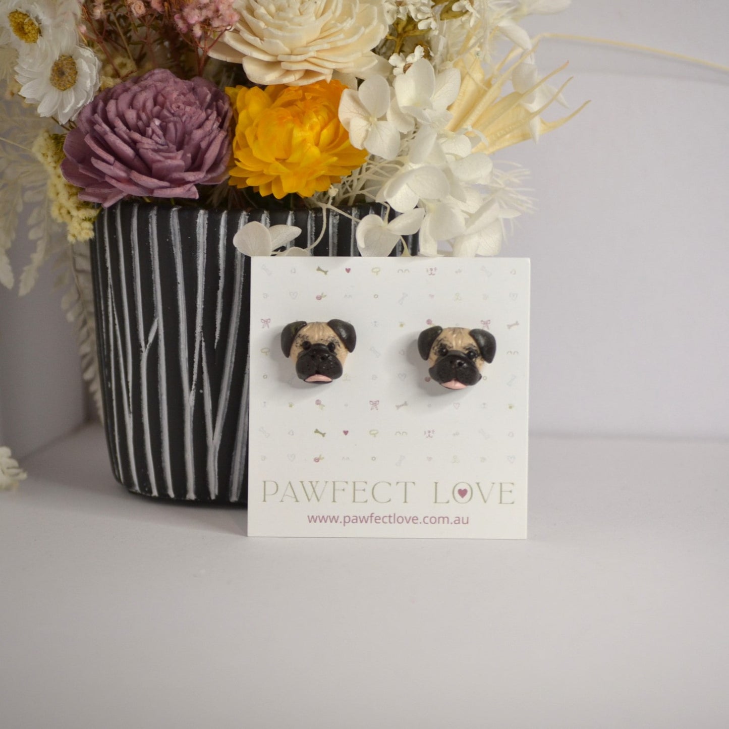 Handmade Pug stud earrings by Pawfect Love, positioned in front of dried flower arrangement